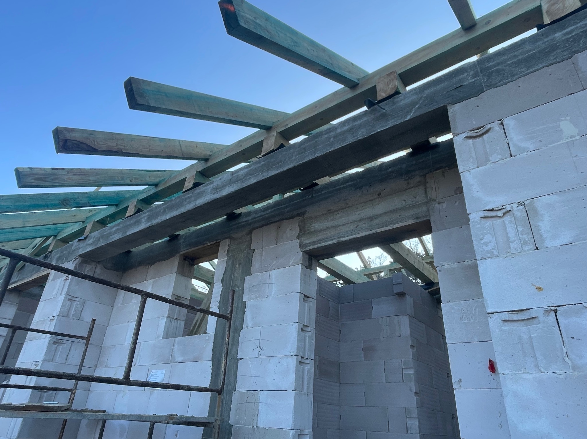 Surowy stan budynku w trakcie budowy: ściany z białych bloczków i drewniana konstrukcja dachu na tle błękitnego nieba. Widok z dołu, podkreślający etap prac.