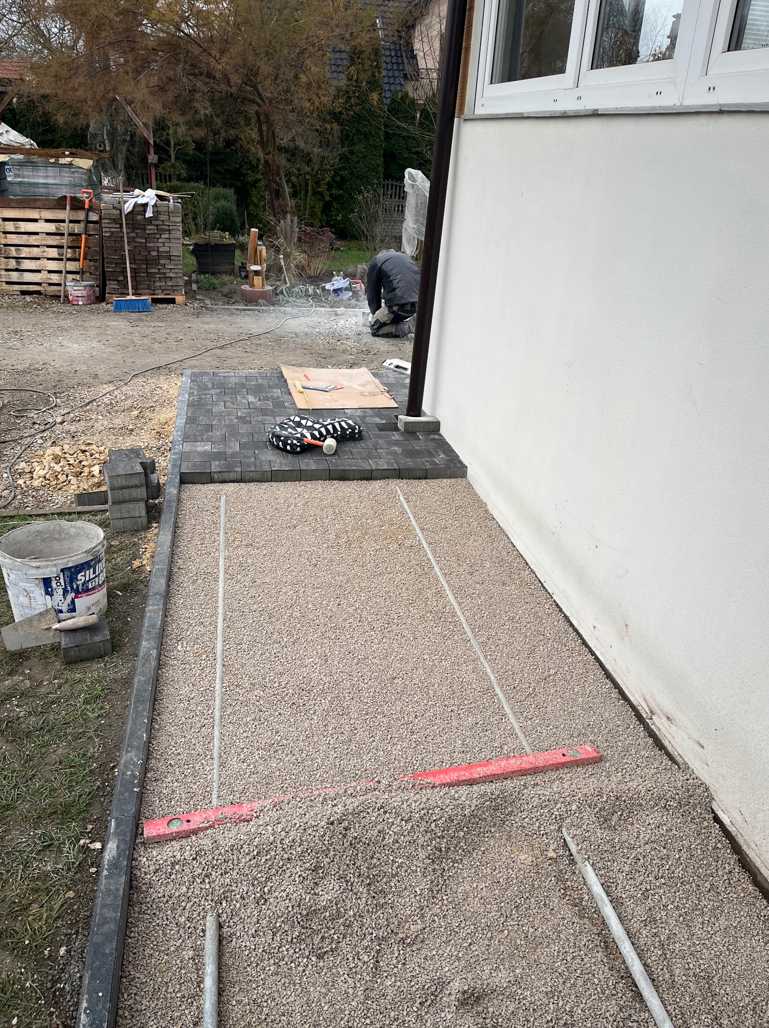 Układanie kostki brukowej przy ścianie budynku. Częściowo ułożona kostka, podsypka z piasku, poziomica i narzędzia. Prace brukarskie w toku, widoczne materiały budowlane.