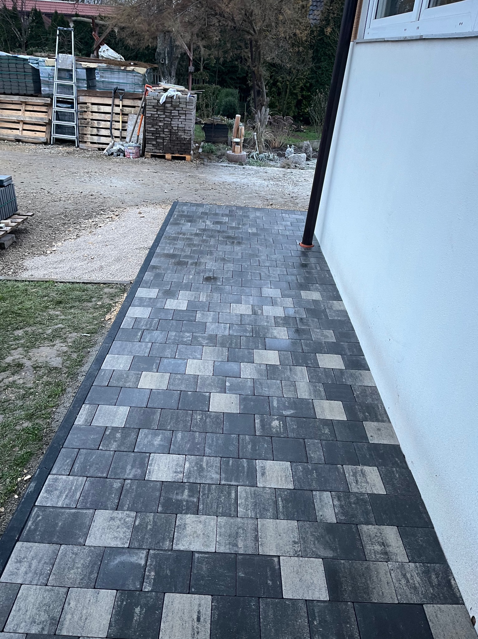Ułożona kostka brukowa w odcieniach szarości tworząca ścieżkę przy białej ścianie budynku. W tle widoczne palety z materiałami budowlanymi i drabina.