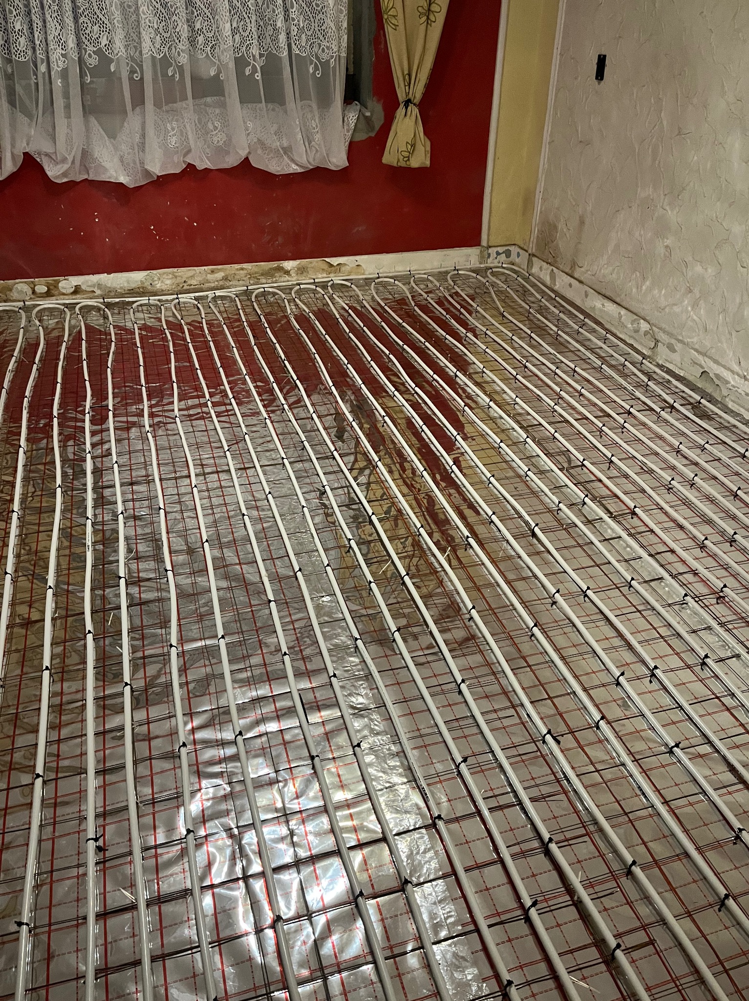 Ułożone rury ogrzewania podłogowego na folii izolacyjnej z siatką, widoczne fragmenty ścian w trakcie remontu, okno z firanką w tle.