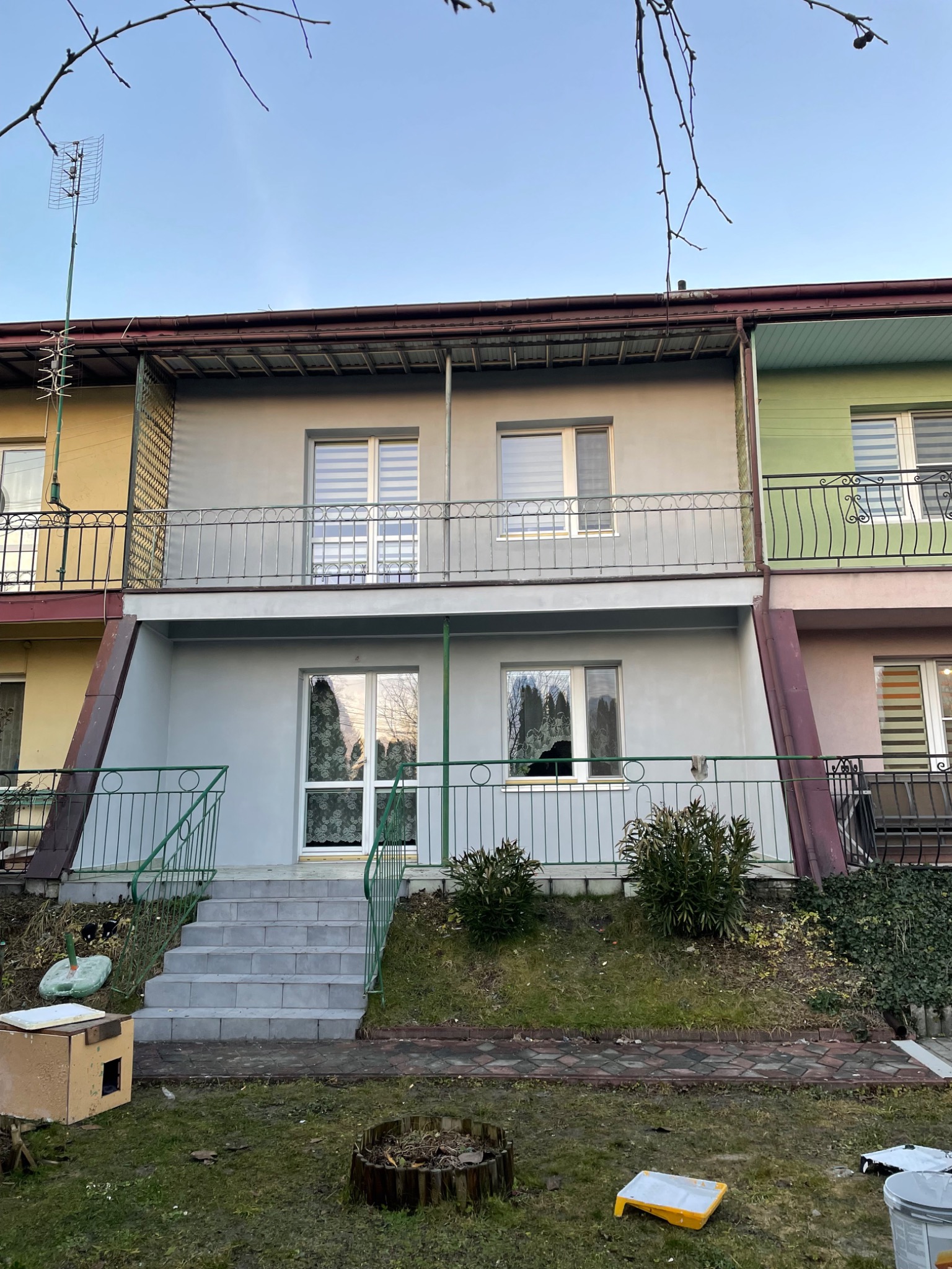 Szary, dwukondygnacyjny segment po remoncie elewacji z zielonymi balustradami i nowymi schodami wejściowymi. Widoczne ślady prac malarskich na trawniku.