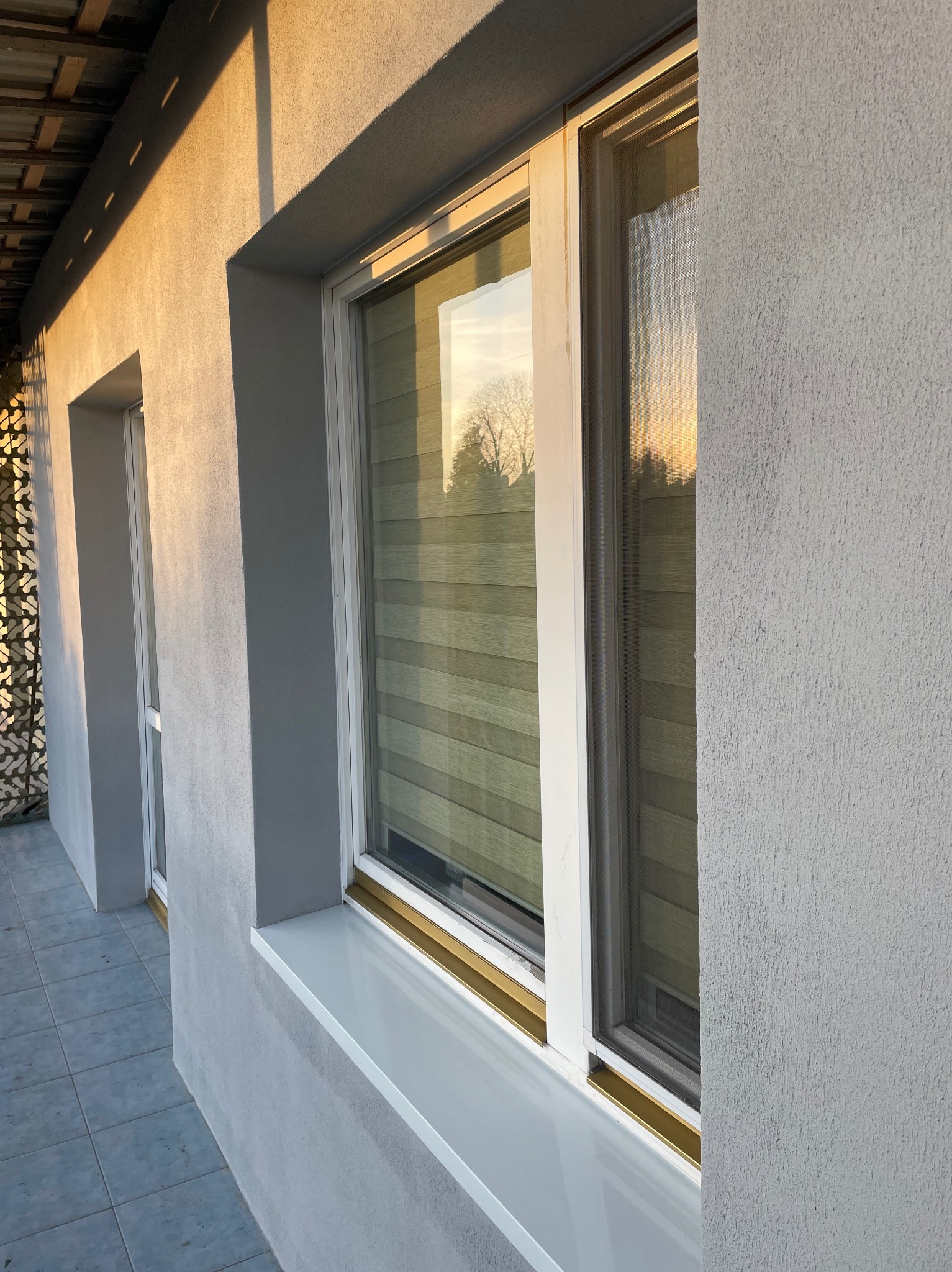 Ściana budynku po malowaniu elewacji z oknami z roletami, widoczne refleksy zachodzącego słońca. Szara, chropowata faktura ściany. Ujęcie z dołu, lekko z boku.