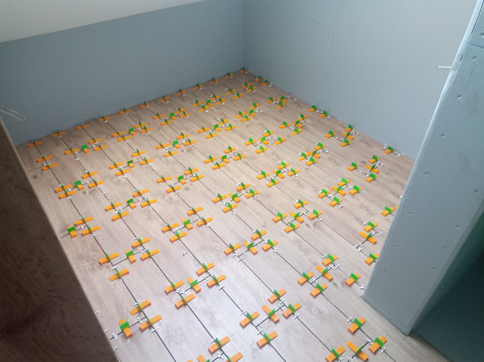 Układanie paneli podłogowych z użyciem klipsów poziomujących w jasnym pomieszczeniu. Widoczne panele imitujące drewno i szare ściany. Ujęcie z góry.