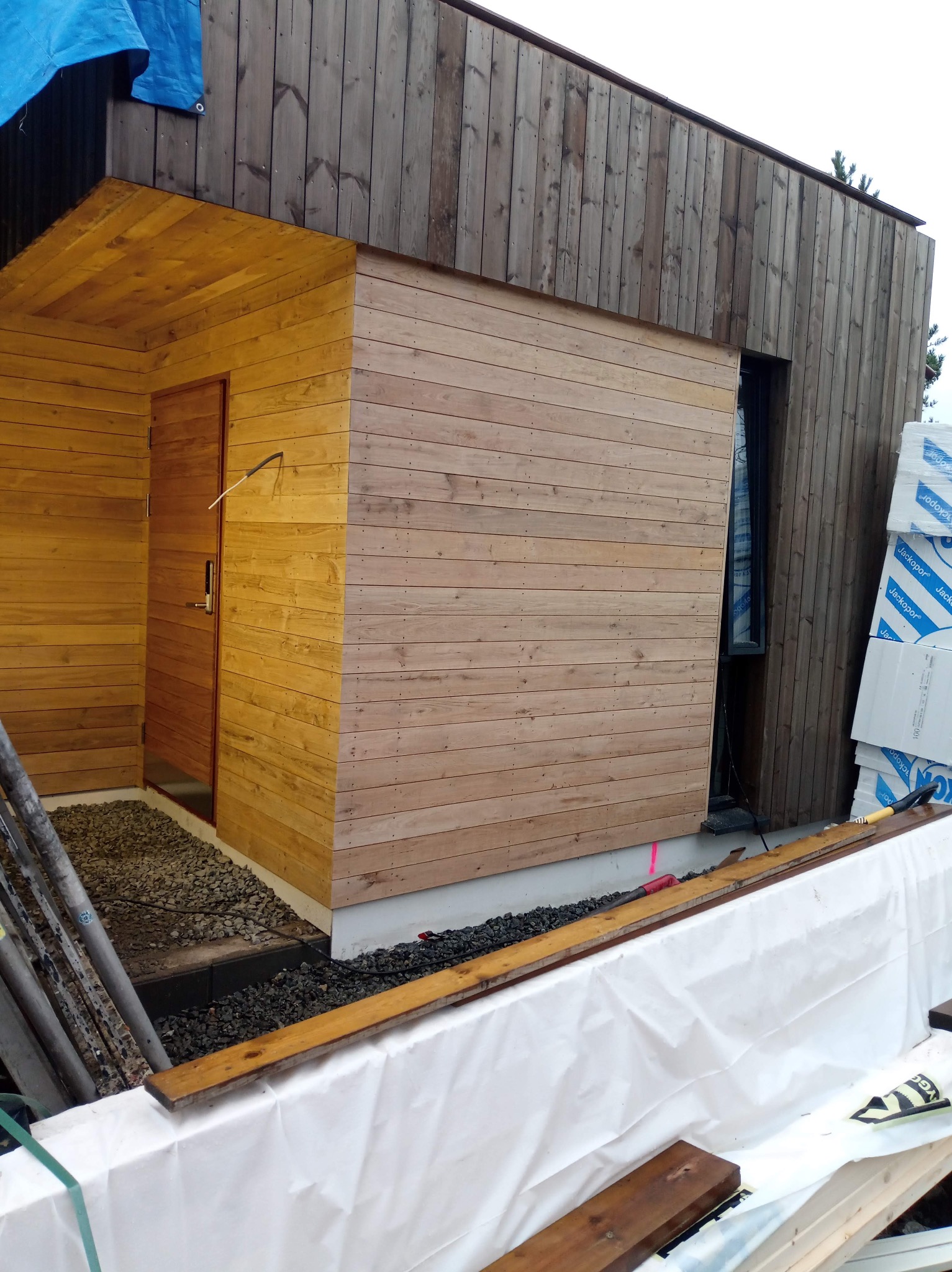 Drewniana elewacja domu w trakcie montażu, widoczne deski w dwóch odcieniach brązu, okno i wejście. Częściowo zakryte materiały budowlane.
