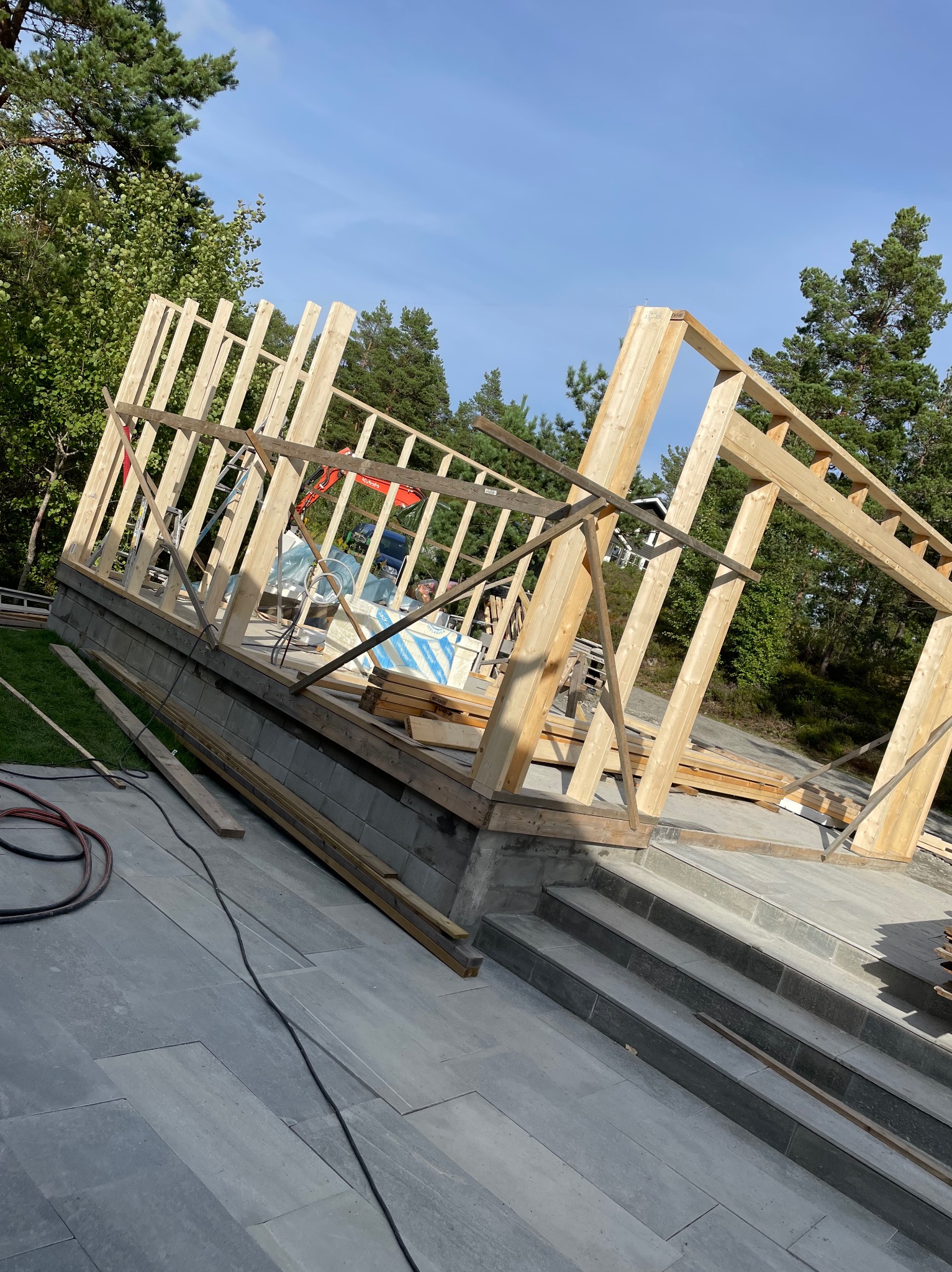 Drewniana konstrukcja szkieletowa budynku w trakcie budowy, widoczna na tle zieleni i błękitnego nieba. Elementy konstrukcyjne z jasnego drewna, fundament z szarej kostki.