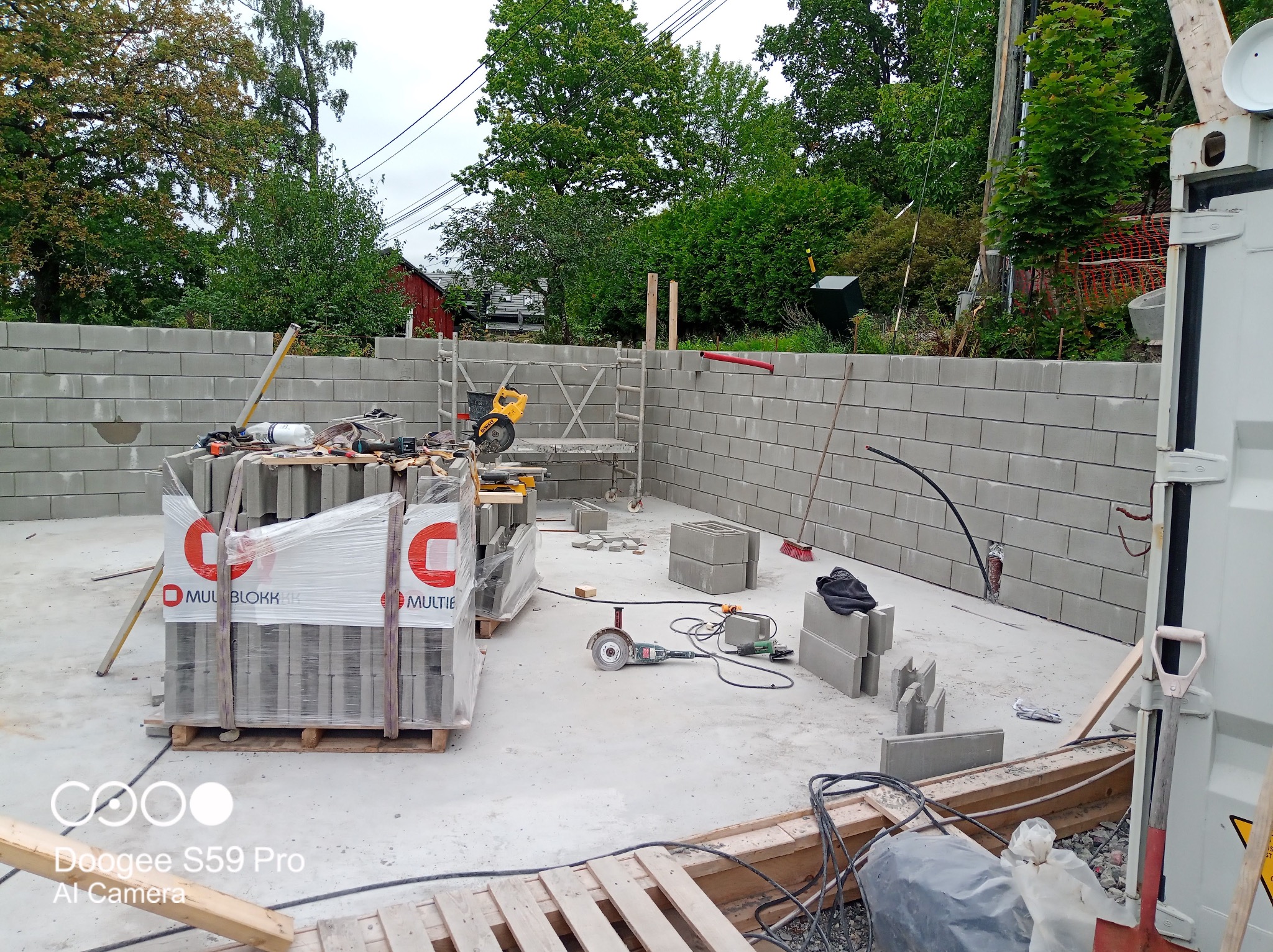 Wznoszenie ściany z szarych bloczków betonowych Multiblokk. Widoczne narzędzia murarskie, paleta z bloczkami i fragment krajobrazu z zielenią w tle.