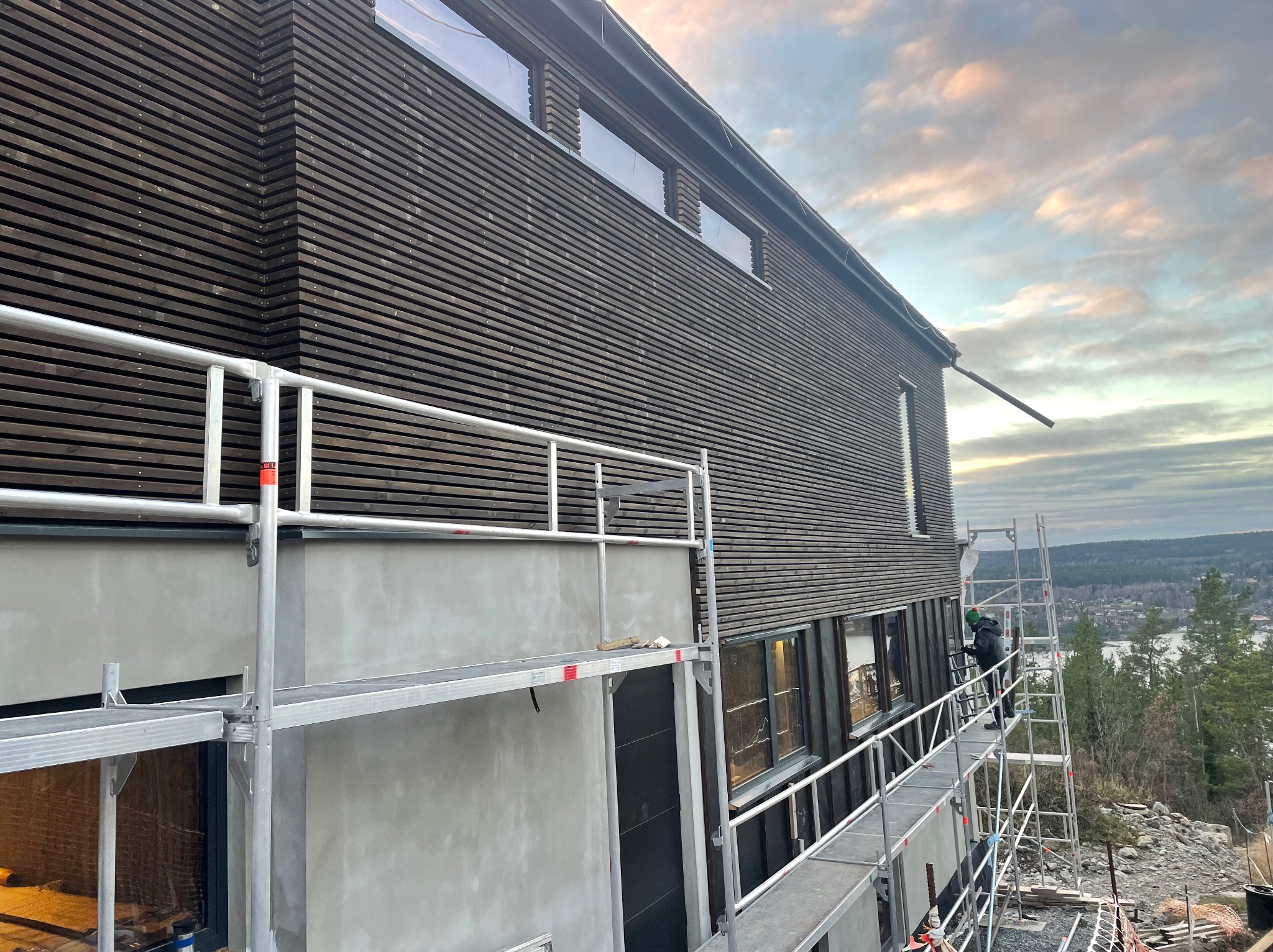 Elewacja domu z ciemnego drewna, częściowo otynkowana, z rusztowaniem. Widoczny pracownik na rusztowaniu i krajobraz w tle. Ujęcie z dołu, w pochmurny dzień.