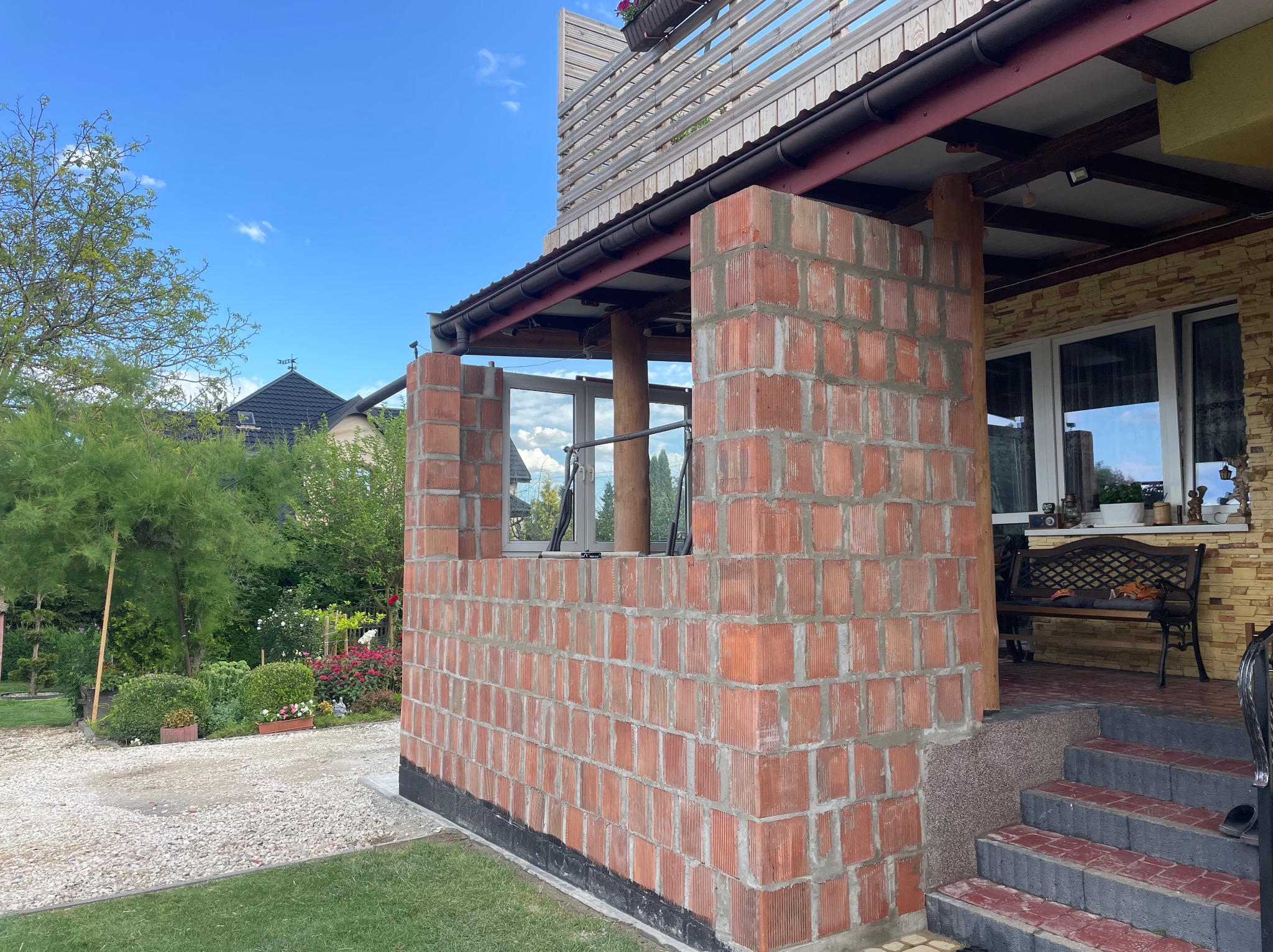 Murowana ściana z czerwonej cegły na zewnątrz budynku w trakcie budowy, widoczne okno i fragment dachu. Prace murarskie w toku, świeża zaprawa.