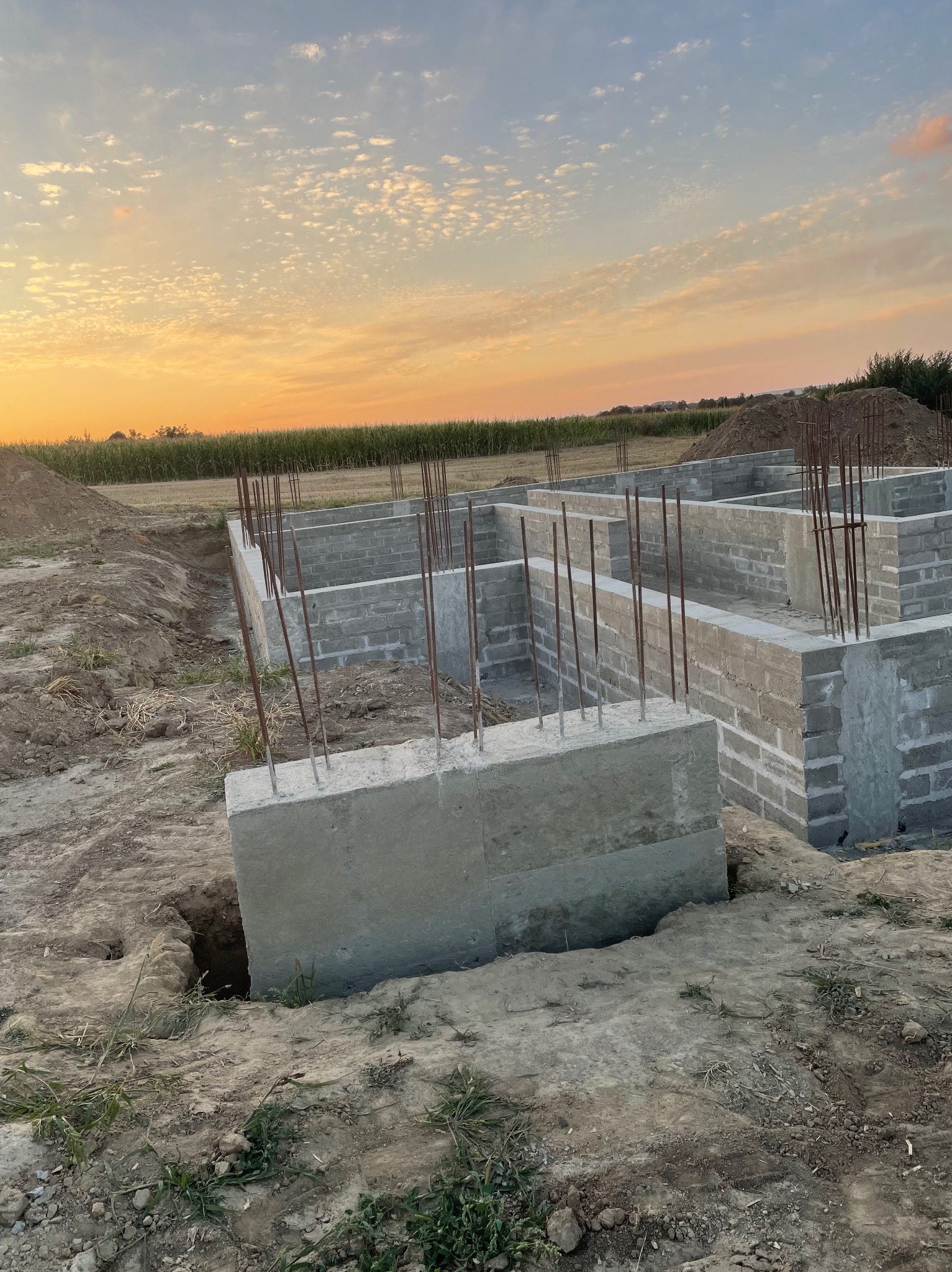 Wykopane fundamenty z betonowymi bloczkami i wystającymi prętami zbrojeniowymi na tle pola i zachodzącego słońca. Etap budowy domu.