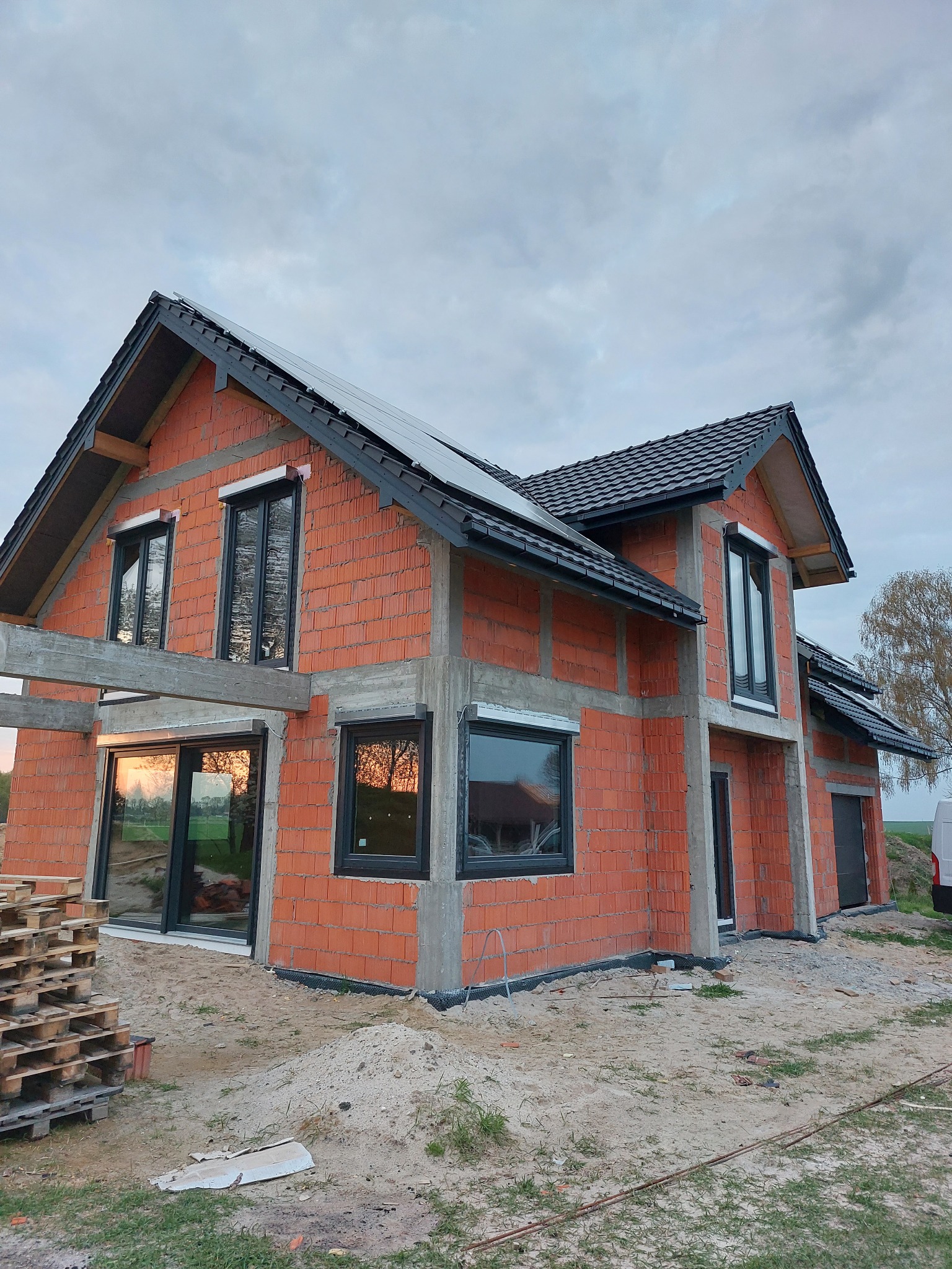 Surowy stan domu z czerwonej cegły z czarnymi oknami i roletami zewnętrznymi, widoczne palety i piasek, dach pokryty dachówką, panele słoneczne na dachu.