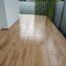 KAJA Grzegorz Dajnowski - Wykończony balkon z drewnopodobną terakotą, szarą balustradą i zieloną ścianą z roślin. Donica z fioletowymi kwiatami dodaje koloru.