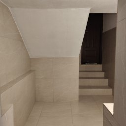 KAJA Grzegorz Dajnowski - Wykończone kafelkami schody i ściany w neutralnych barwach, prowadzące do ciemnych drzwi. Widoczne detale wykończenia i oświetlenie.