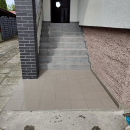 KAJA Grzegorz Dajnowski - Wykończone płytkami schody wejściowe z kolumną z cegły klinkierowej. Widoczna osoba w drzwiach. Jasne płytki na schodach i podłodze.