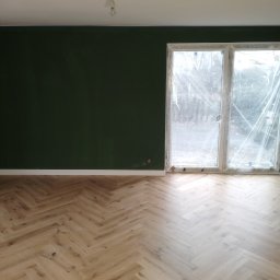 KAJA Grzegorz Dajnowski - Pokój po remoncie: parkiet w jodełkę, ściany pomalowane na ciemnozielono, okno zabezpieczone folią malarską, białe listwy przypodłogowe.