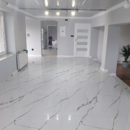 KAJA Grzegorz Dajnowski - Jasne, przestronne wnętrze po remoncie: białe marmurowe płytki podłogowe z brązowymi żyłami, szare ściany, białe drzwi i sufit z drewnianymi belkami. Nowoczesne oświetlenie.