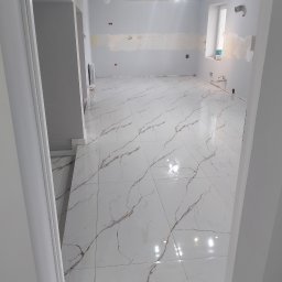 KAJA Grzegorz Dajnowski - Wykończone wnętrze z błyszczącą, białą podłogą imitującą marmur z brązowymi żyłami. Ściany szare, częściowo przygotowane do malowania. Widok zza futryny.