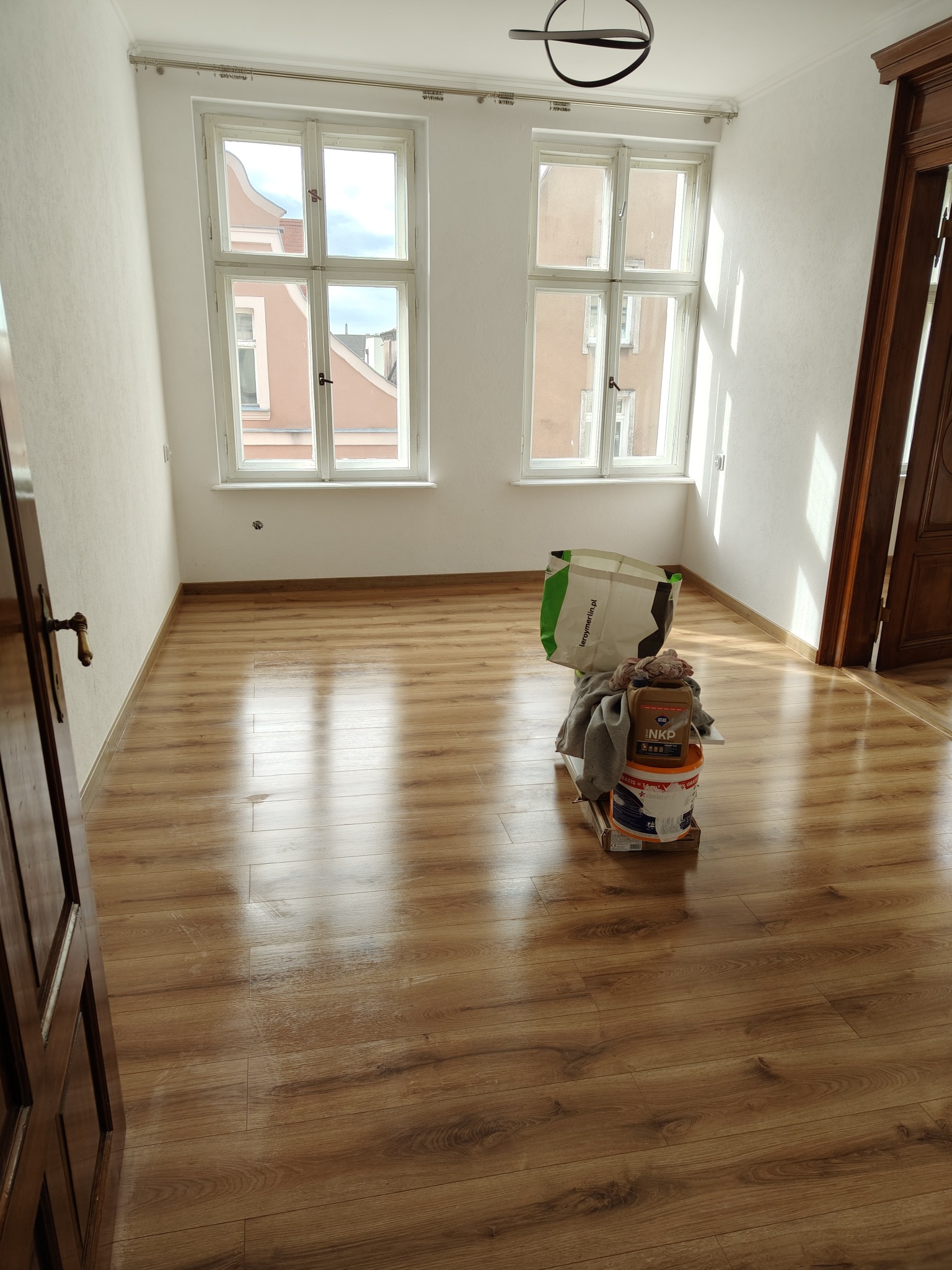 Jasne wnętrze z drewnianą podłogą, dwa okna z widokiem na budynki. Na podłodze wiadro z farbą i torba, sugerujące przygotowania do malowania.