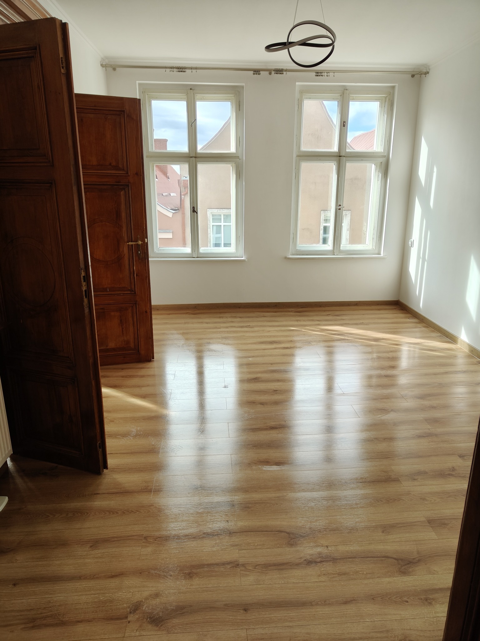 Jasne wnętrze po remoncie: drewniana podłoga, białe ściany, okna z widokiem na budynki. Otwarta, ciemna, drewniana stolarka drzwiowa. Nowoczesna lampa sufitowa.