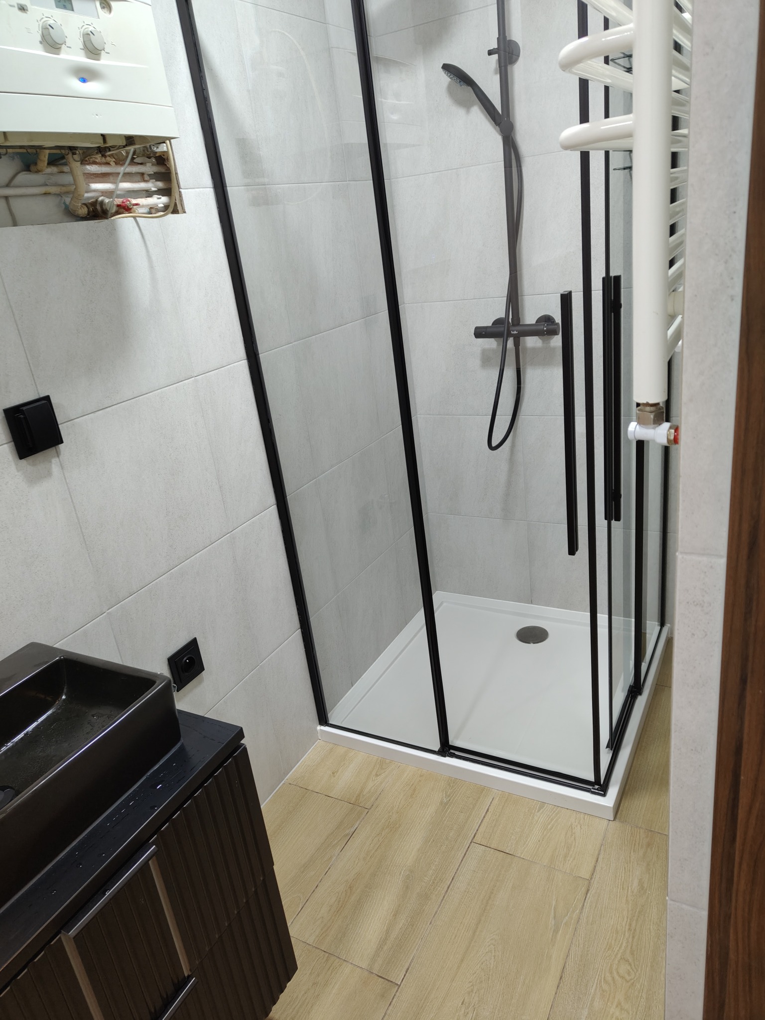 Nowoczesna łazienka po remoncie: kabina prysznicowa z czarnymi ramami, umywalka na szafce w kolorze grafitu, jasne płytki ścienne i podłogowe imitujące drewno.