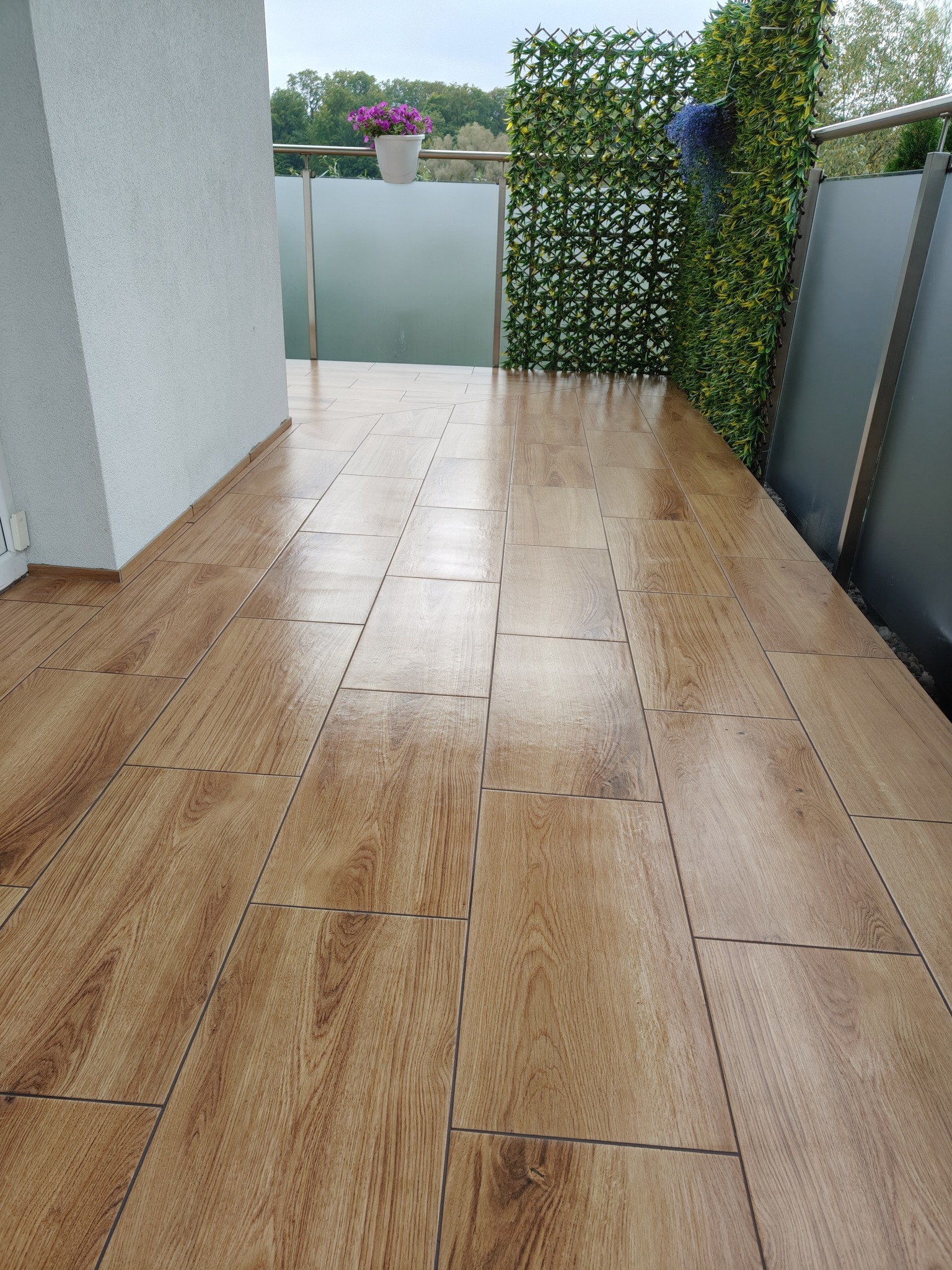 Wykończony balkon z drewnopodobną terakotą, szarą balustradą i zieloną ścianą z roślin. Donica z fioletowymi kwiatami dodaje koloru.