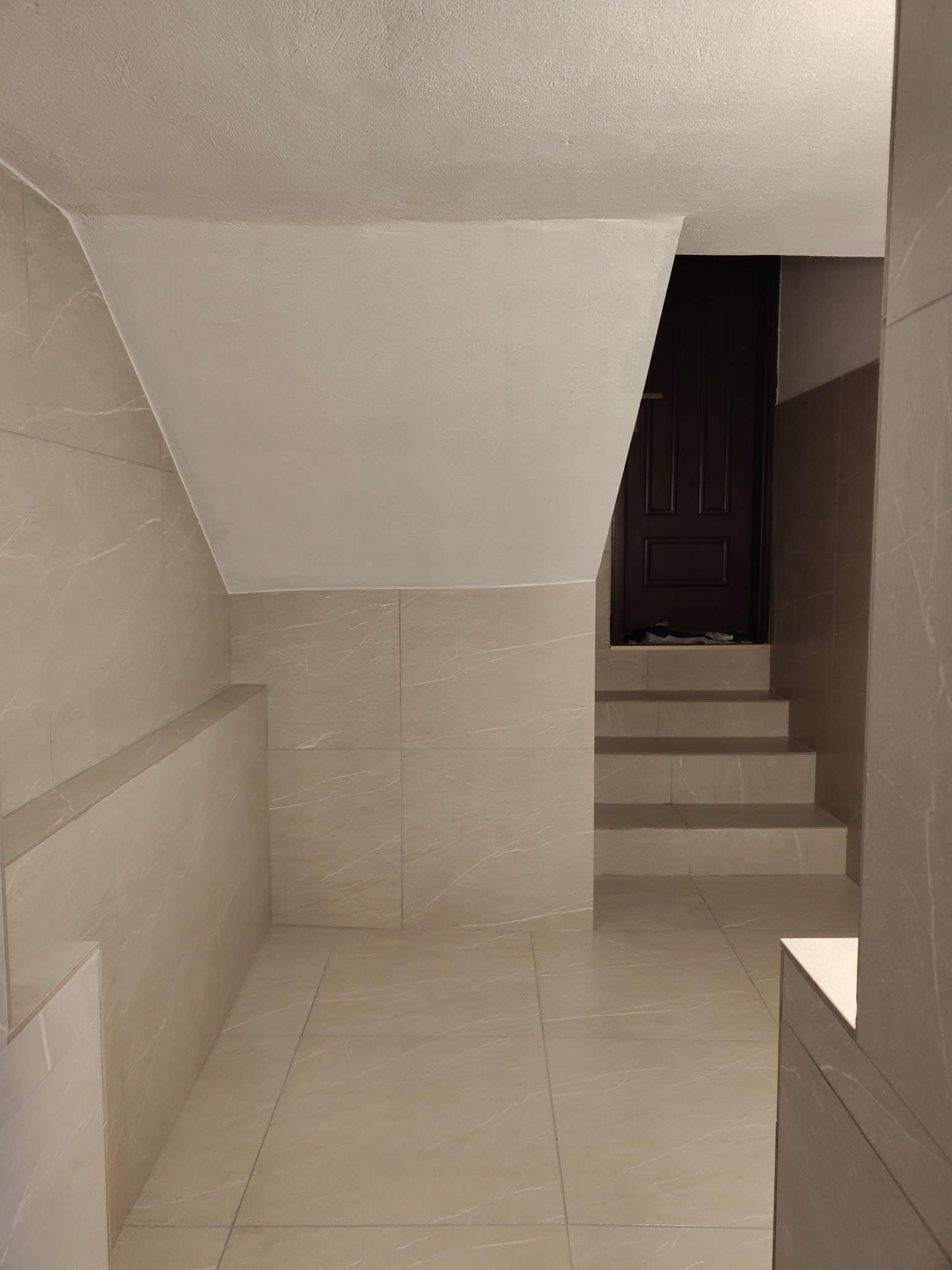 Wykończone kafelkami schody i ściany w neutralnych barwach, prowadzące do ciemnych drzwi. Widoczne detale wykończenia i oświetlenie.