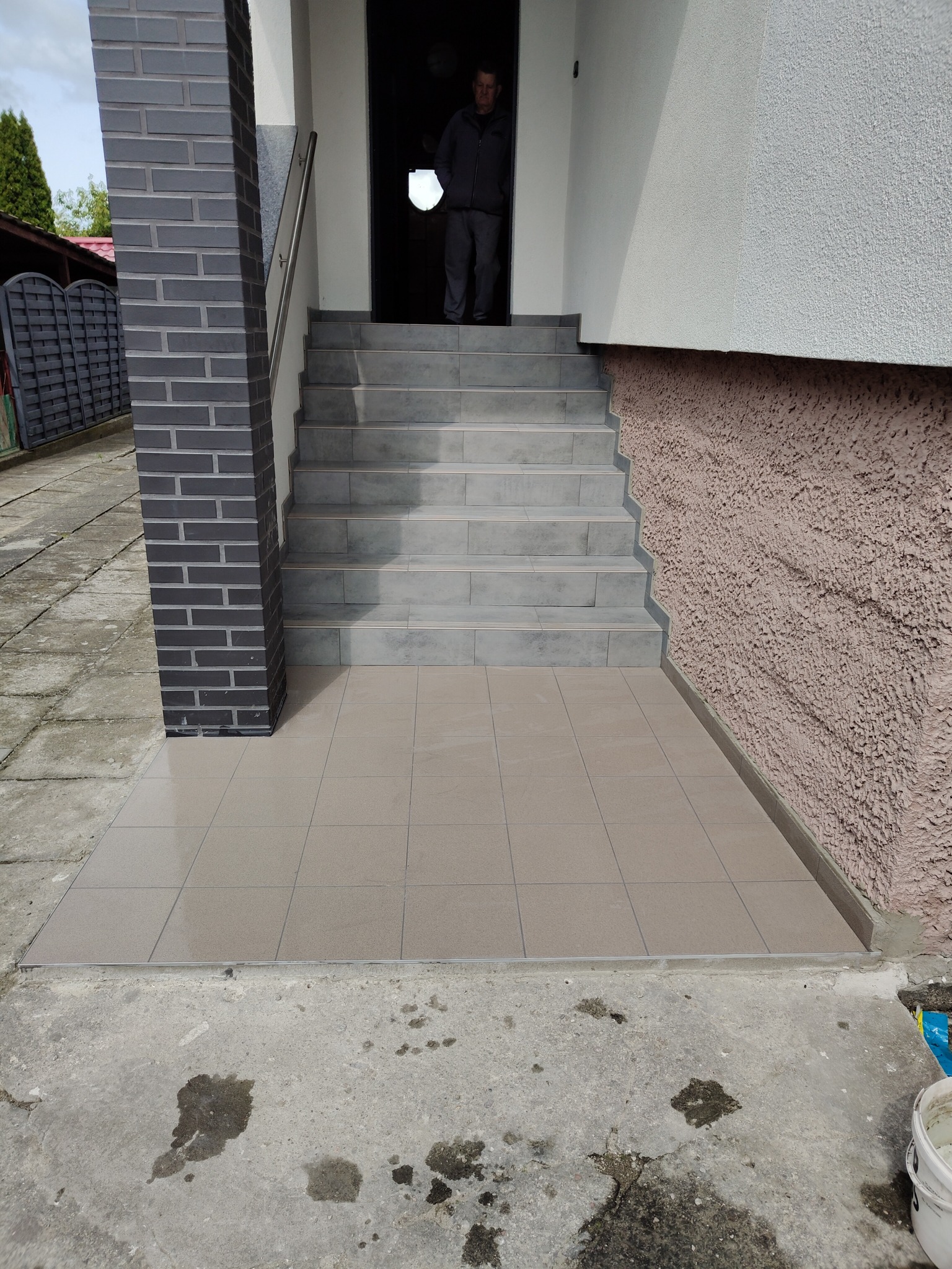 Wykończone płytkami schody wejściowe z kolumną z cegły klinkierowej. Widoczna osoba w drzwiach. Jasne płytki na schodach i podłodze.