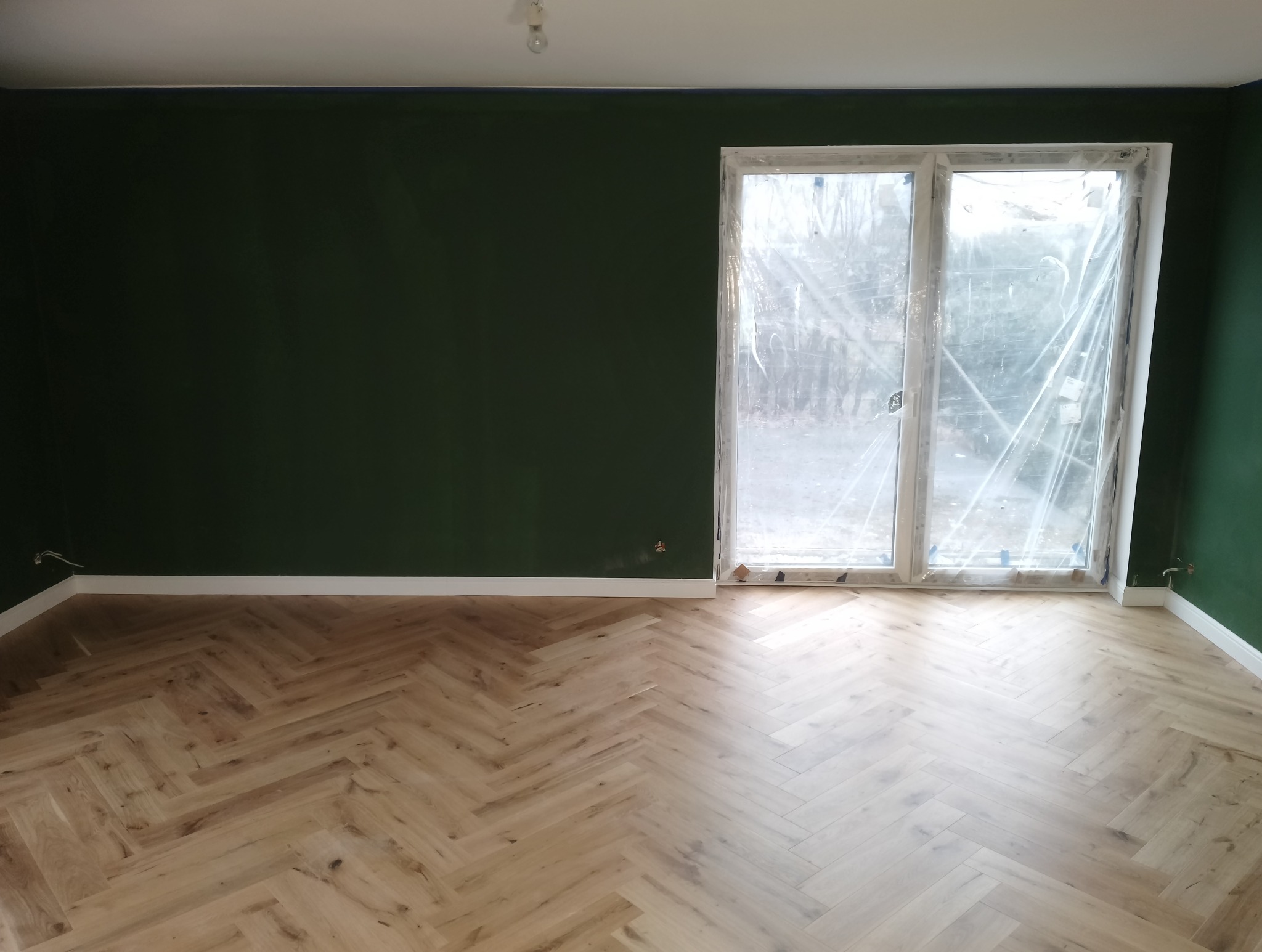 Pokój po remoncie: parkiet w jodełkę, ściany pomalowane na ciemnozielono, okno zabezpieczone folią malarską, białe listwy przypodłogowe.