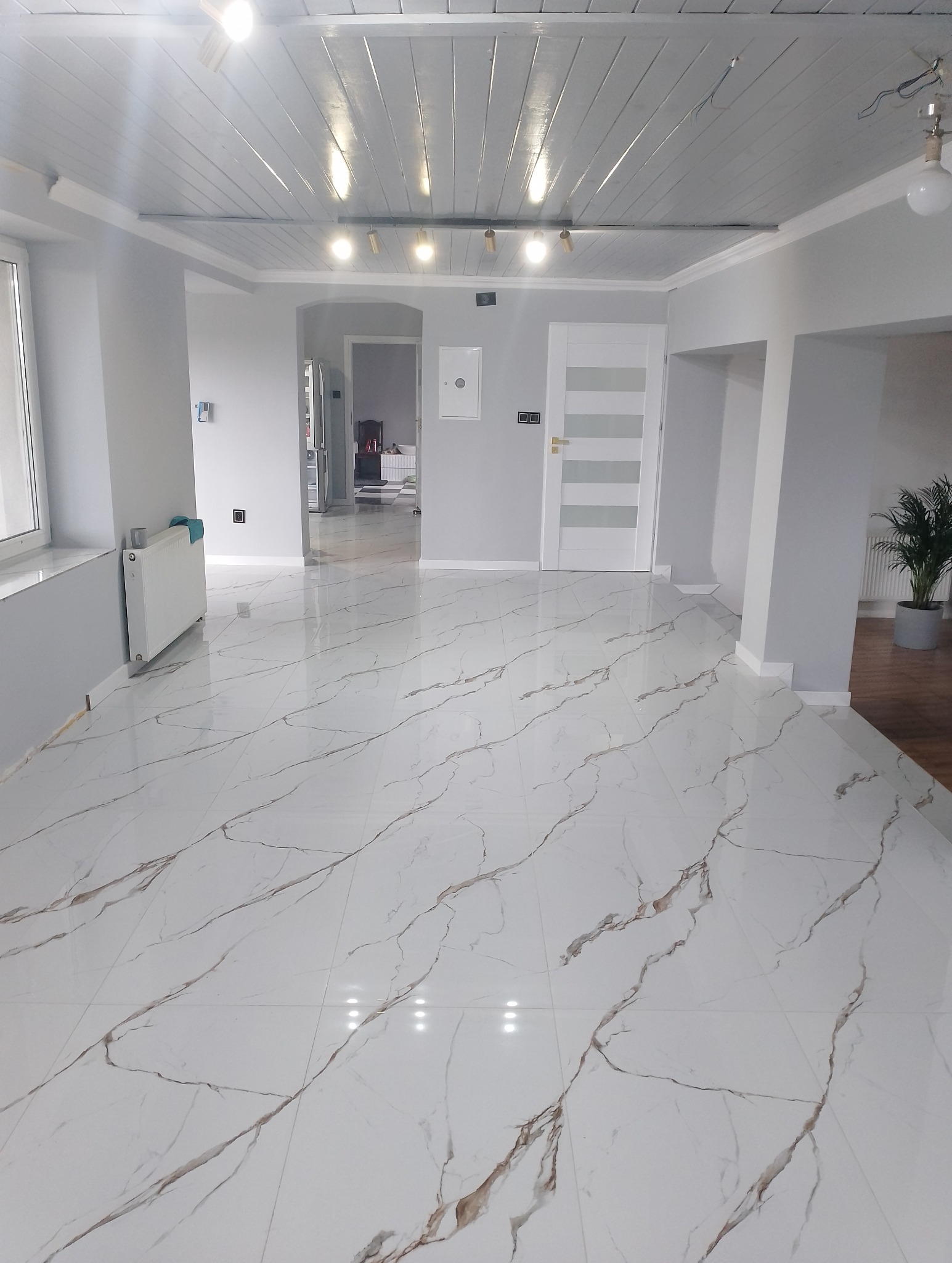 Jasne, przestronne wnętrze po remoncie: białe marmurowe płytki podłogowe z brązowymi żyłami, szare ściany, białe drzwi i sufit z drewnianymi belkami. Nowoczesne oświetlenie.