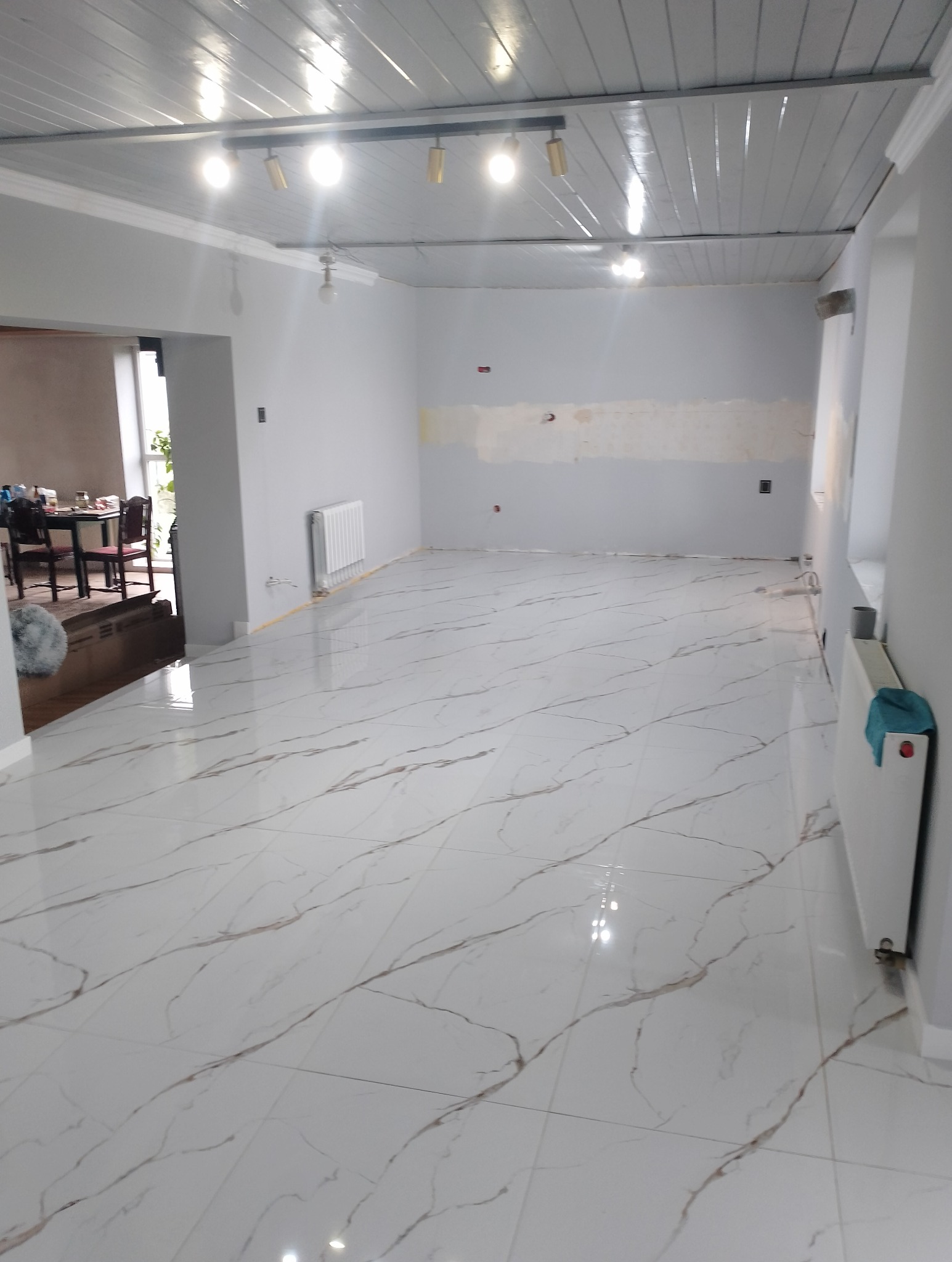Jasne, przestronne wnętrze po remoncie: białe płytki podłogowe z marmurkowym wzorem, szare ściany z fragmentem po szpachlowaniu, biały sufit z oświetleniem.