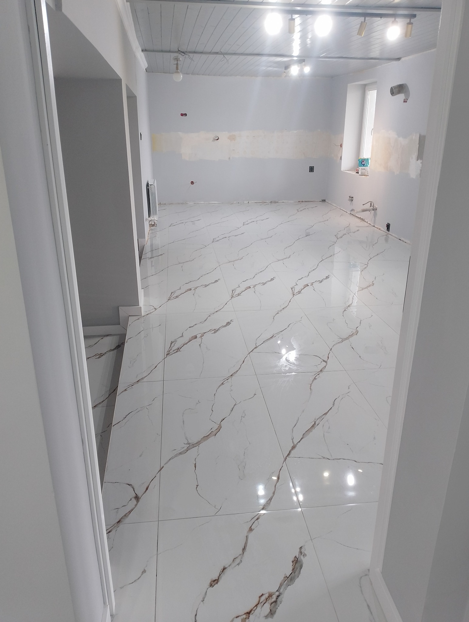 Wykończone wnętrze z błyszczącą, białą podłogą imitującą marmur z brązowymi żyłami. Ściany szare, częściowo przygotowane do malowania. Widok zza futryny.