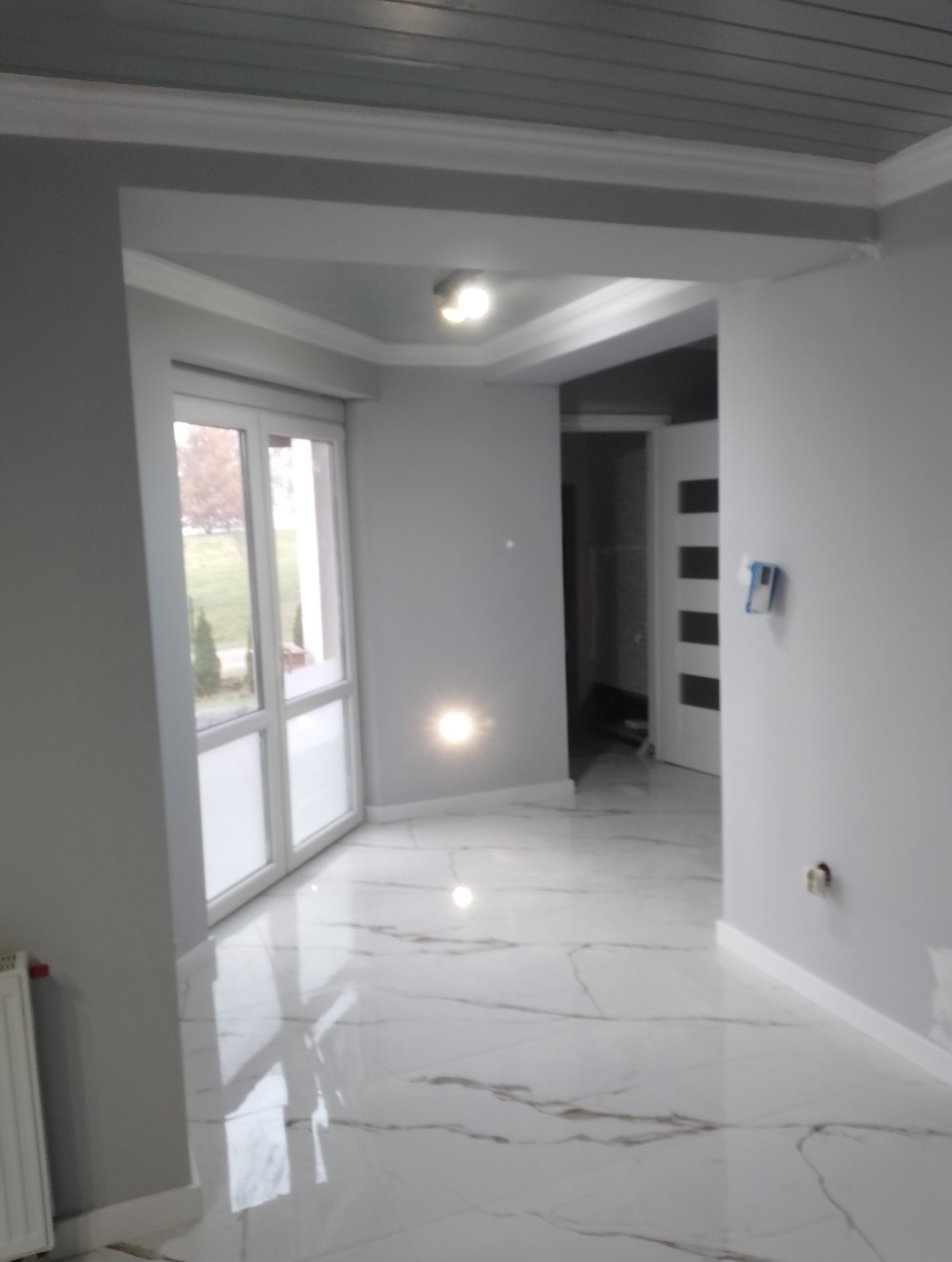 Wykończone wnętrze w szarościach: biała marmurowa podłoga, szare ściany i sufit z drewnianą boazerią. Widoczne okno i drzwi wewnętrzne.