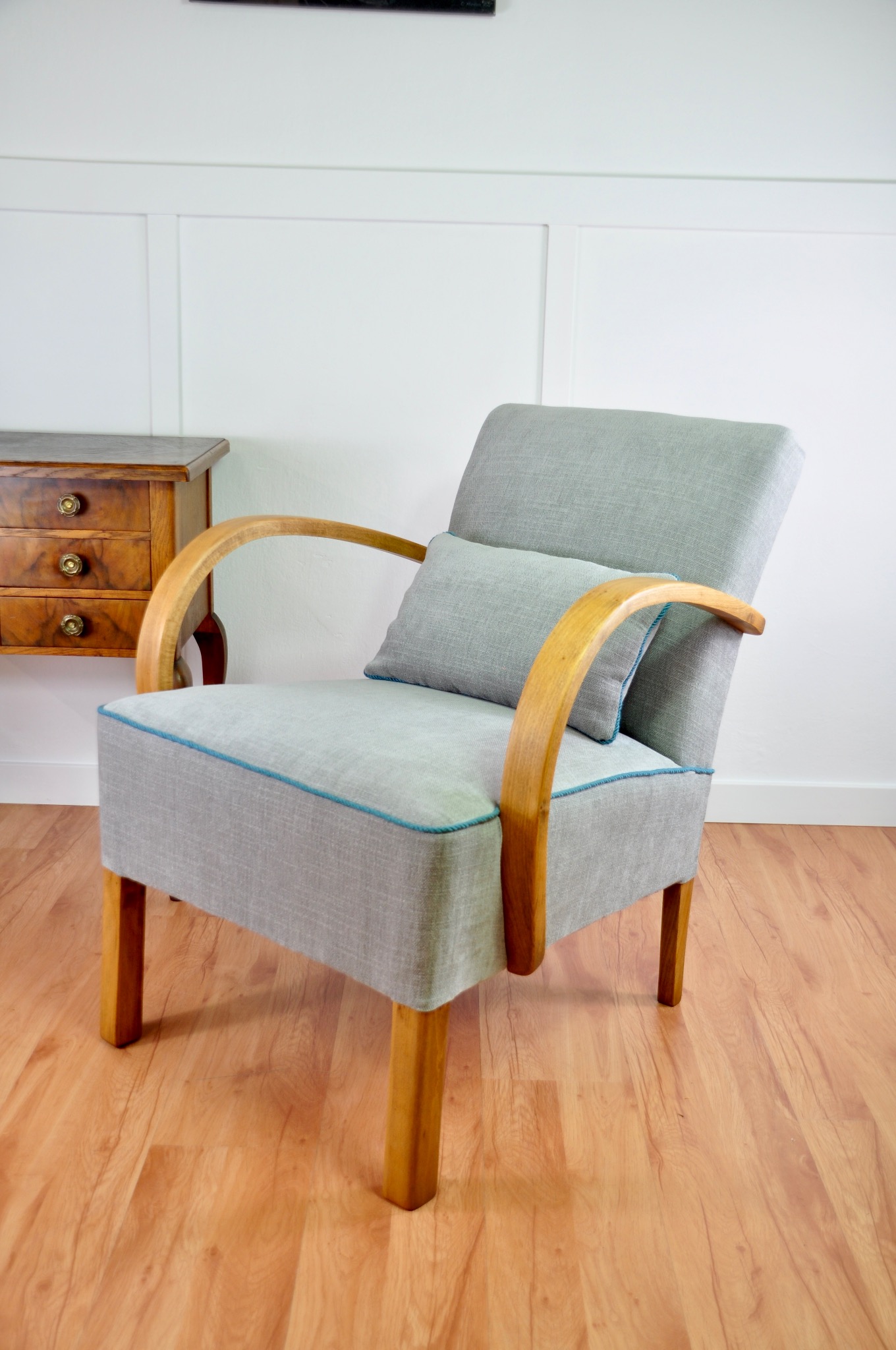 Szary fotel z drewnianymi podłokietnikami i niebieską lamówką, uzupełniony poduszką. W tle komoda i biała ściana z panelami. Ujęcie w jasnym wnętrzu.