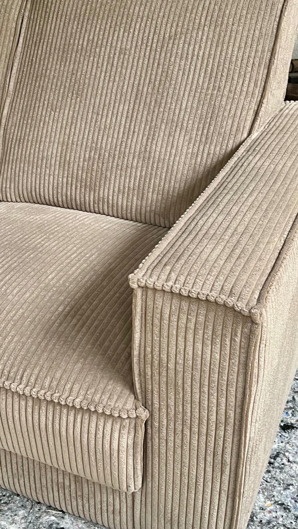 Beżowy, prążkowany fotel z bliska, ukazujący fakturę materiału i detale wykończenia podłokietnika. Widoczny fragment podłogi z resztkami materiału.