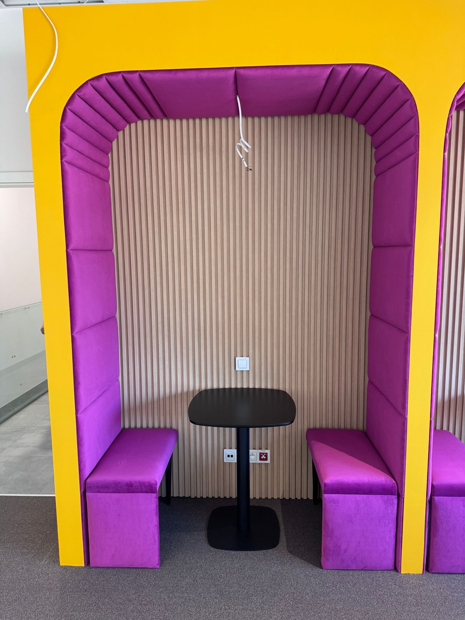 Fioletowa, tapicerowana kabina biurowa z żółtymi elementami, wyposażona w czarny stolik. Pionowe panele ścienne w tle. Nowoczesny design.