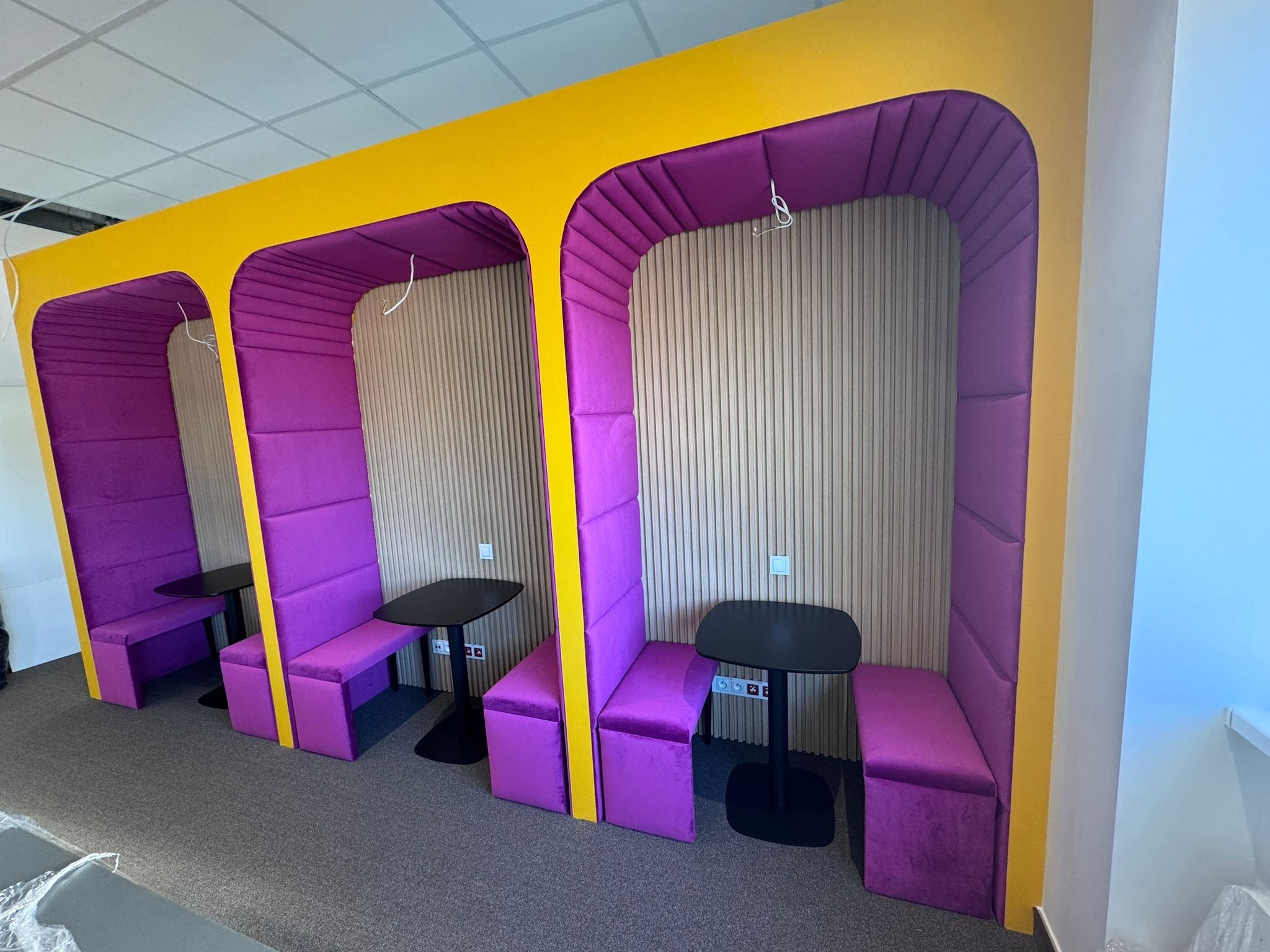 Trzy fioletowe, tapicerowane kabiny z żółtymi obramowaniami i czarnymi stolikami, ustawione w rzędzie na szarej podłodze. Ściany wykończone pionowymi panelami.