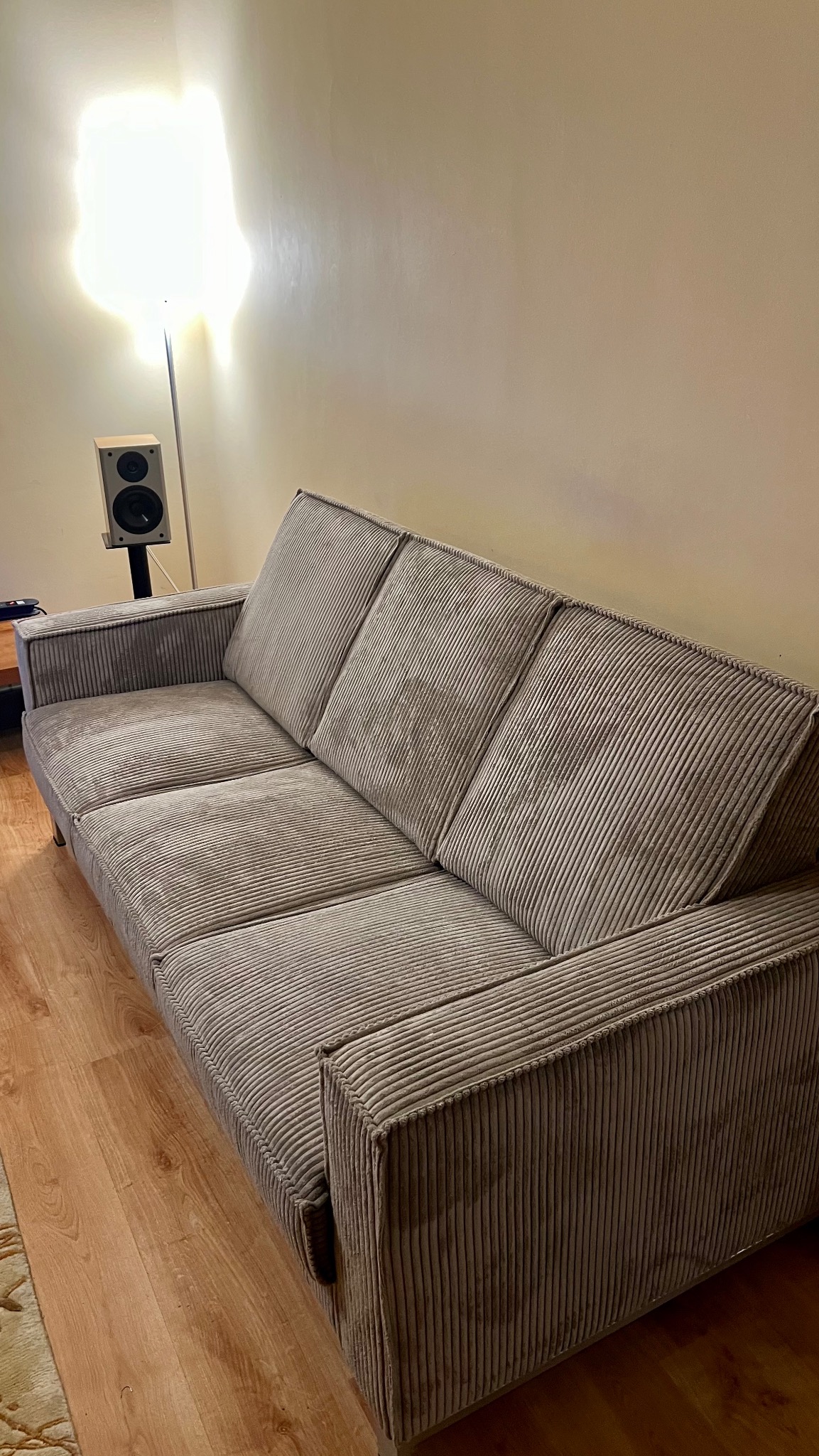 Szary, trzyosobowy, sztruksowy fotel z podłokietnikami w salonie. W tle lampa podłogowa i głośnik. Widok z boku, oświetlenie górne.