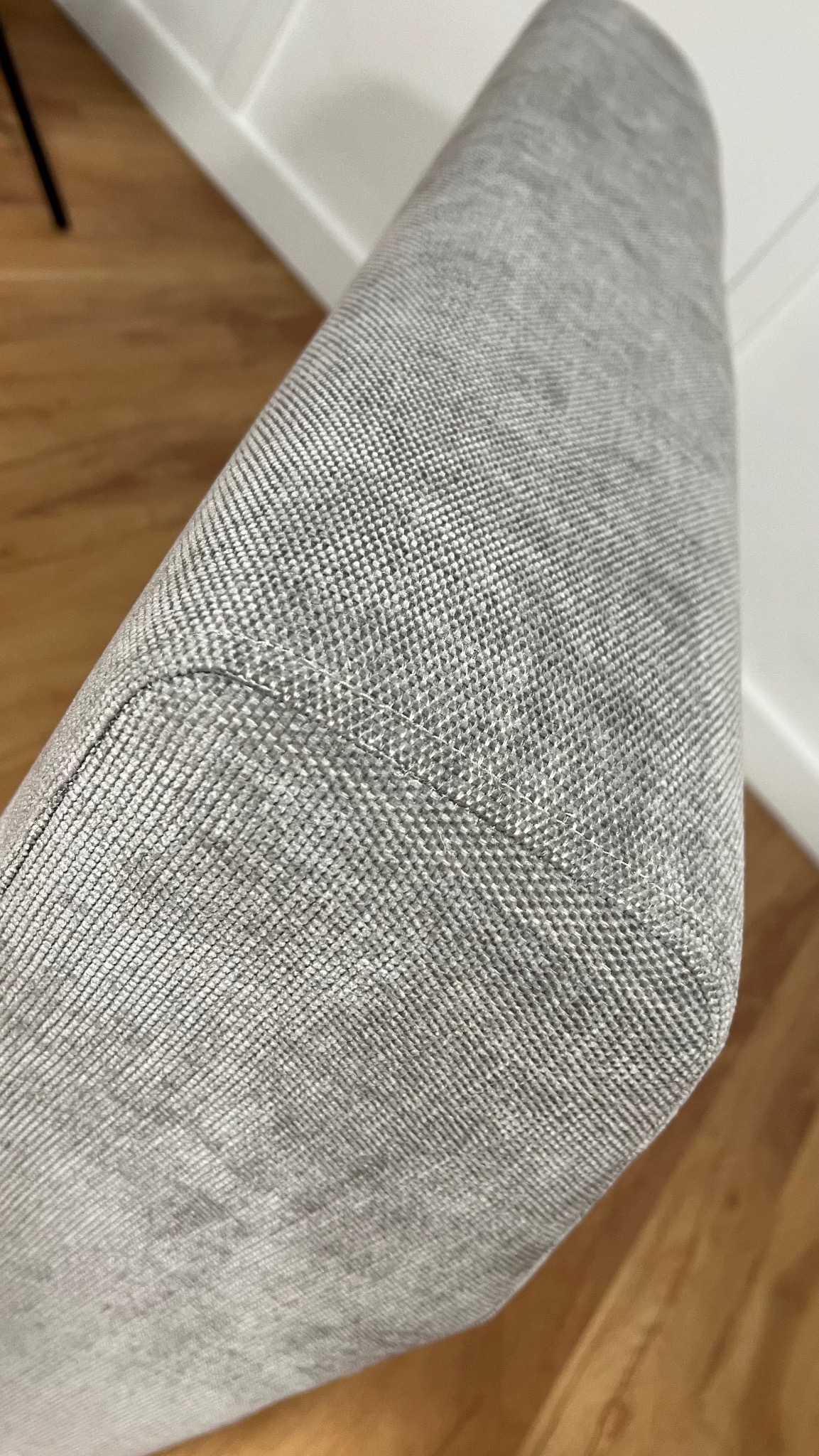 Szary, tapicerowany narożnik krzesła z widocznym szwem, na tle drewnianej podłogi i białej ściany. Detal wykończenia mebla.