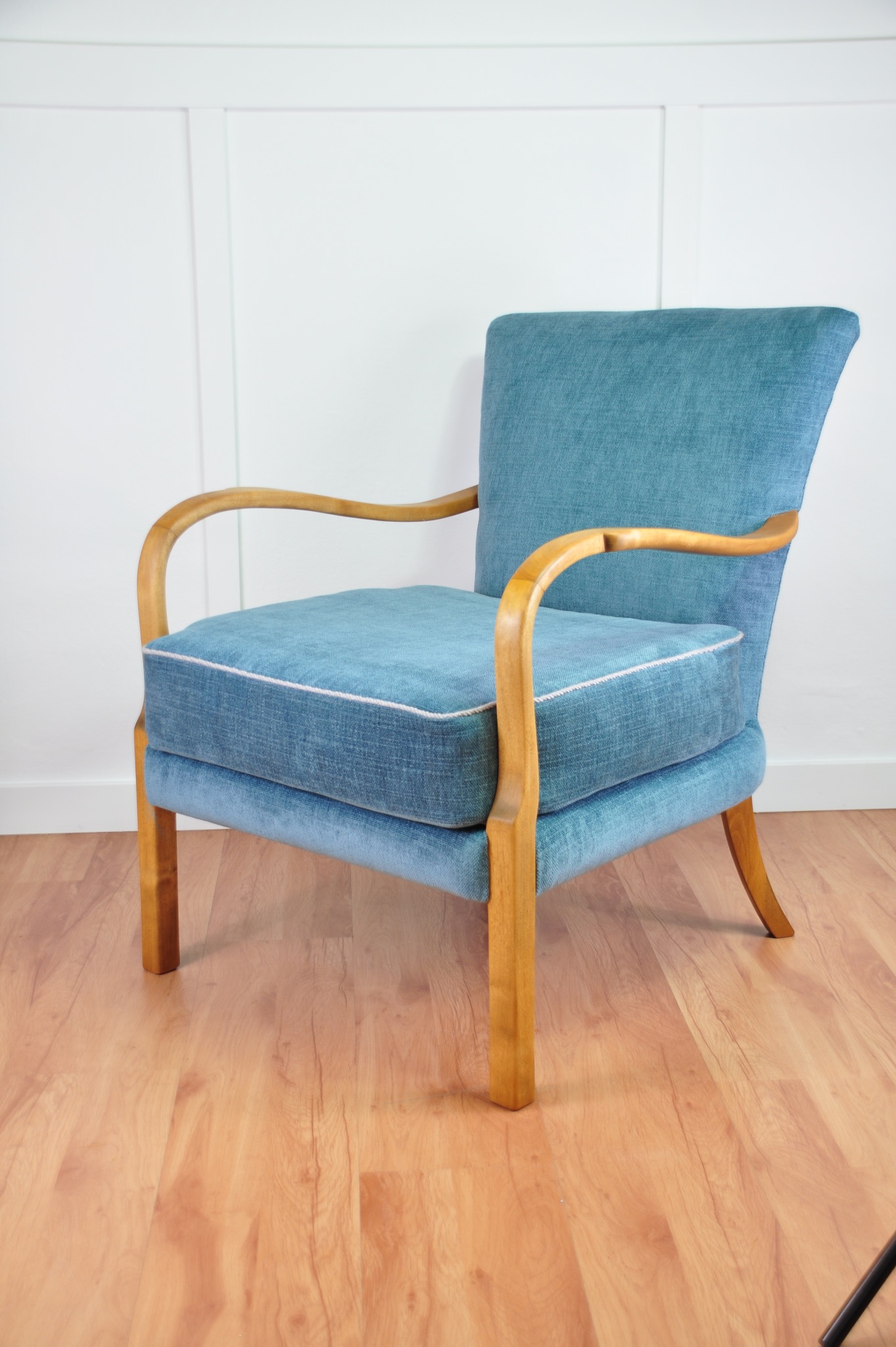 Błękitny fotel z drewnianymi podłokietnikami i lamówką, na parkiecie. Jasne tło z panelami. Fotel po renowacji tapicerskiej, z nową tkaniną i wykończeniem.