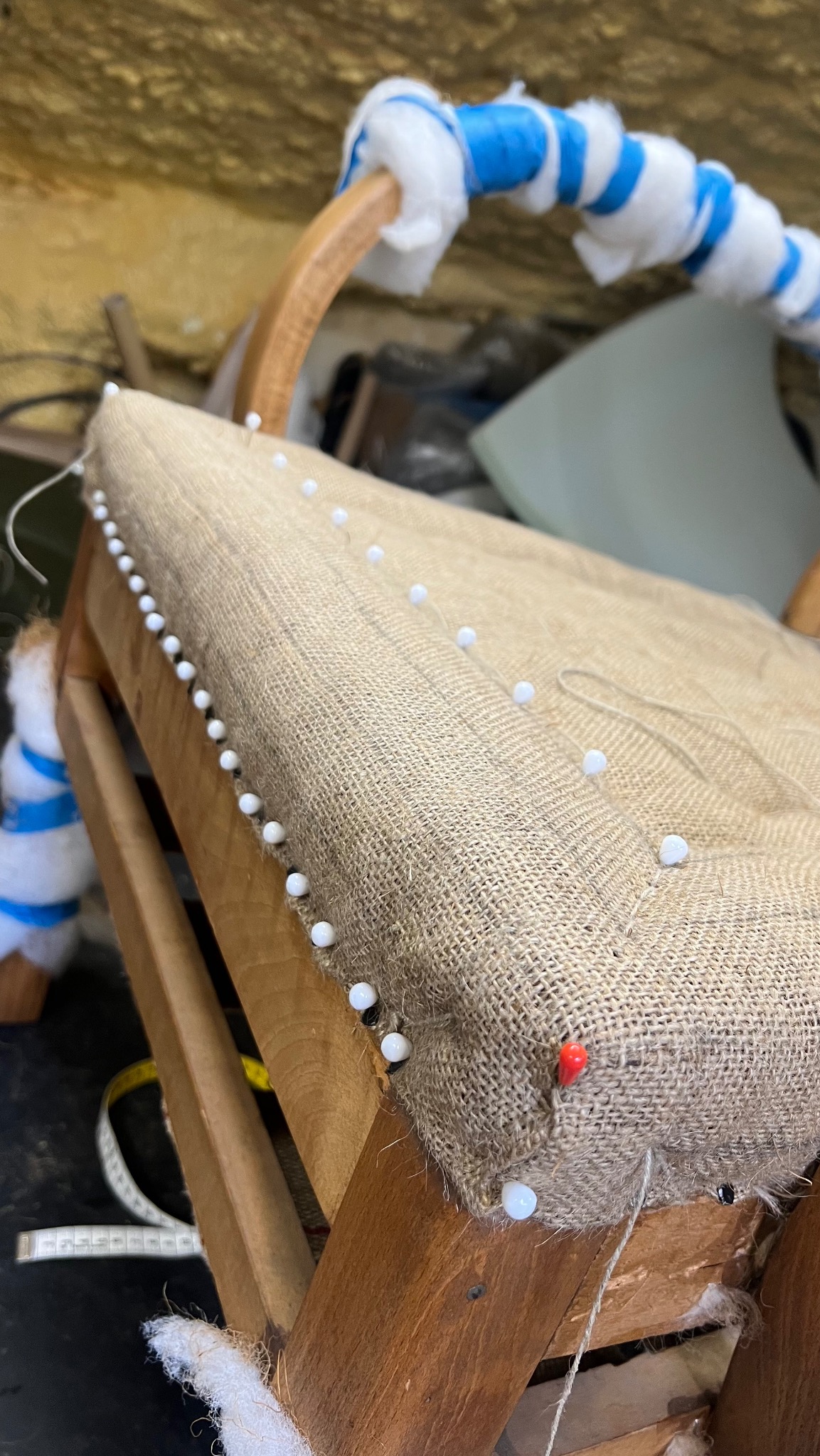 Fragment krzesła w trakcie tapicerowania. Widoczne drewniane elementy, surowa tkanina jutowa z białymi szpilkami i watą. W tle narzędzia tapicerskie.