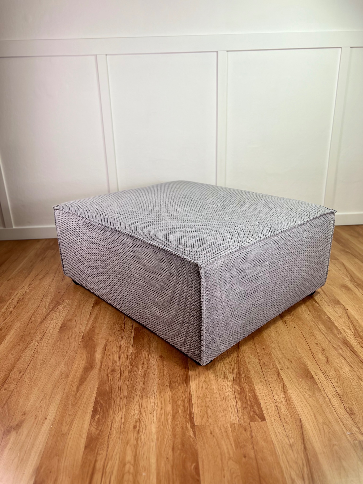 Szary, tapicerowany puf w kształcie sześcianu na drewnianej podłodze, z białym panelem ściennym w tle. Minimalistyczny design, widoczne detale tkaniny.
