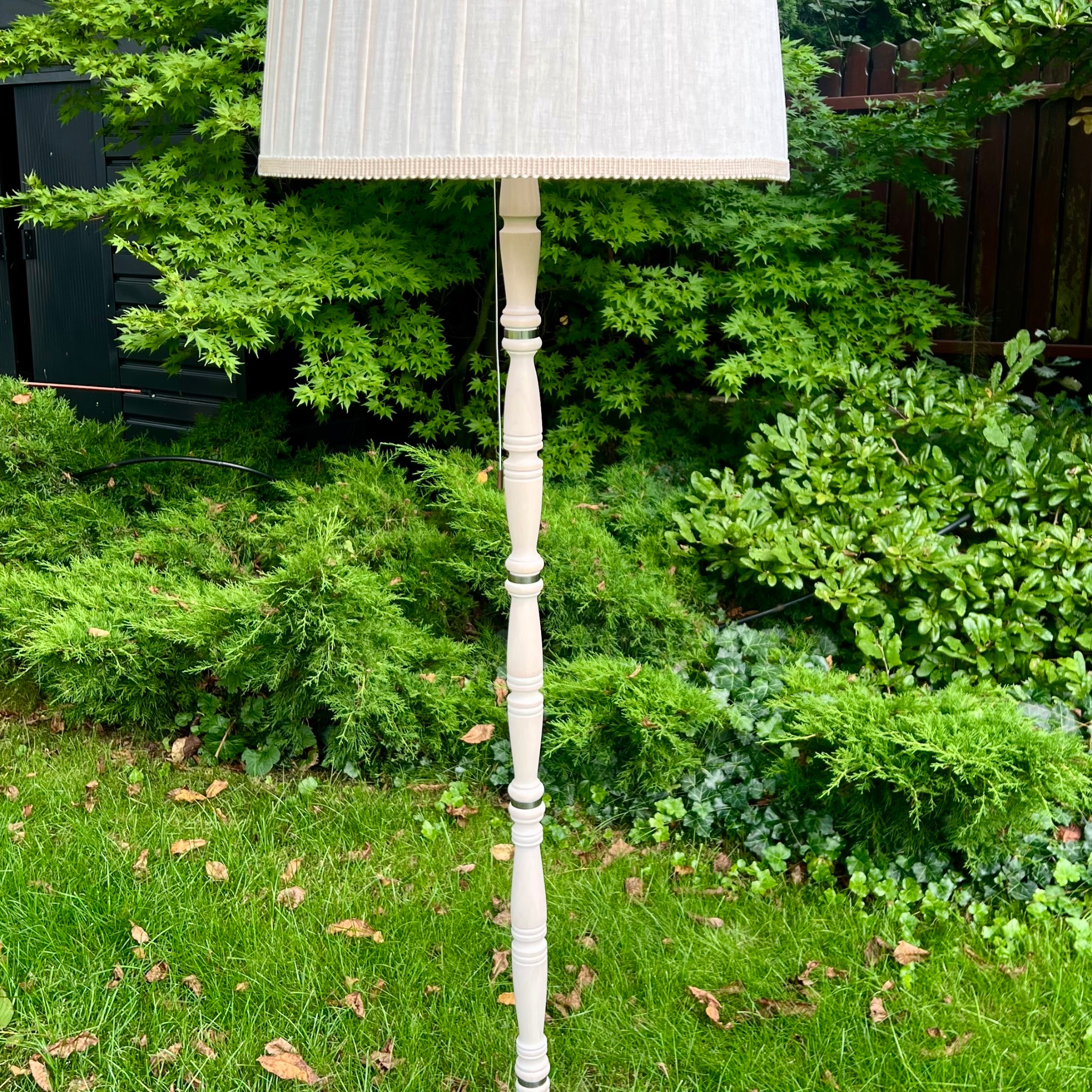 Elegancka, beżowa lampa podłogowa z plisowanym abażurem, ustawiona na trawie przed zielonymi krzewami i ciemną, drewnianą konstrukcją. Dekoracyjny element oświetleniowy.