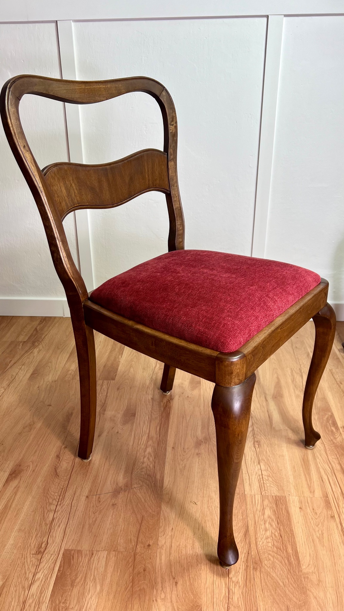 Odrestaurowane krzesło z ciemnego drewna z bordowym siedziskiem na drewnianej podłodze. Widoczne toczone nogi i biała boazeria w tle. Klasyczny styl.