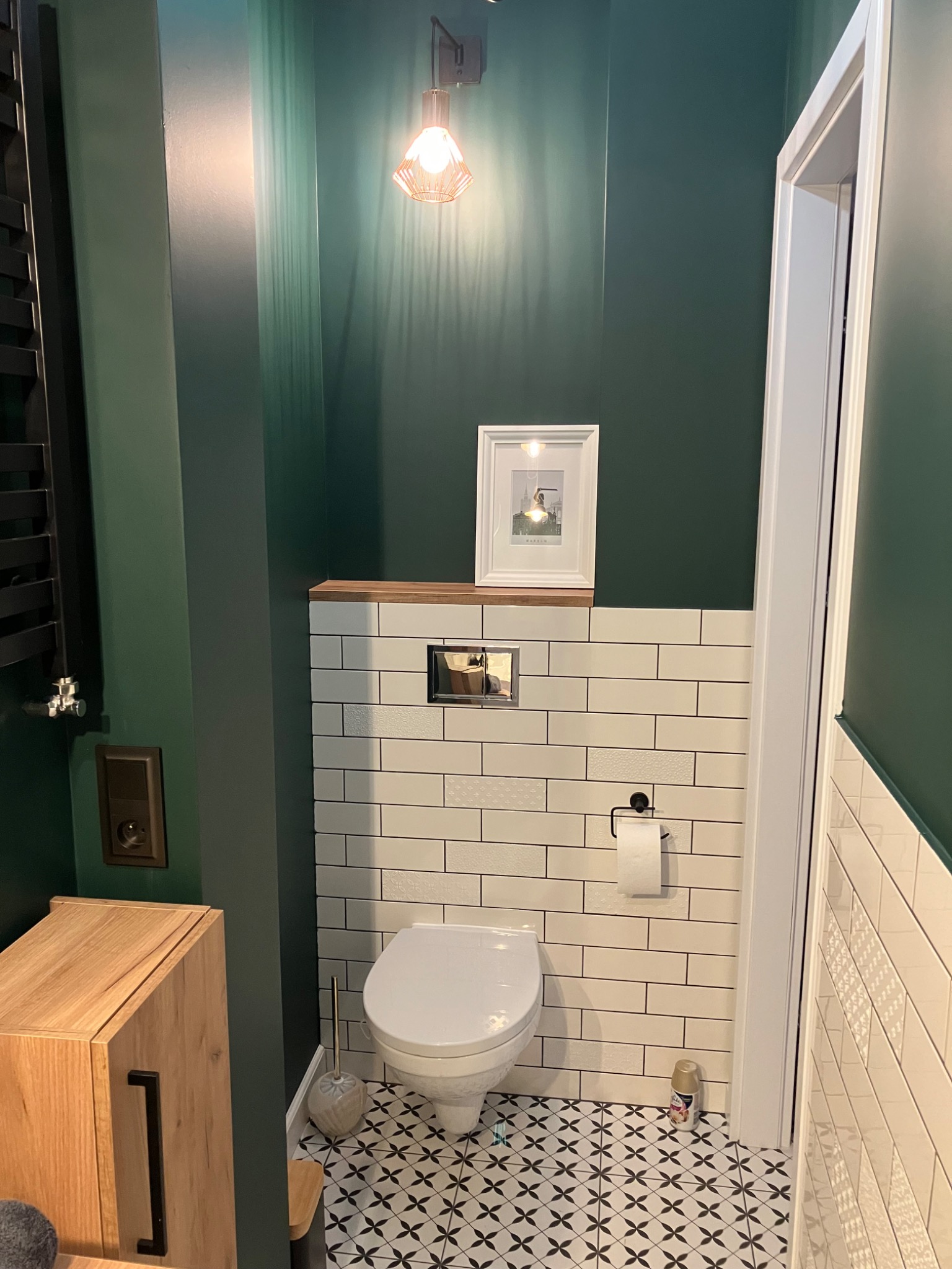 Nowoczesna toaleta w odcieniach zieleni i bieli: geometryczne płytki podłogowe, biała armatura, drewniana szafka i designerska lampa. Minimalistyczny styl z akcentem na detale.