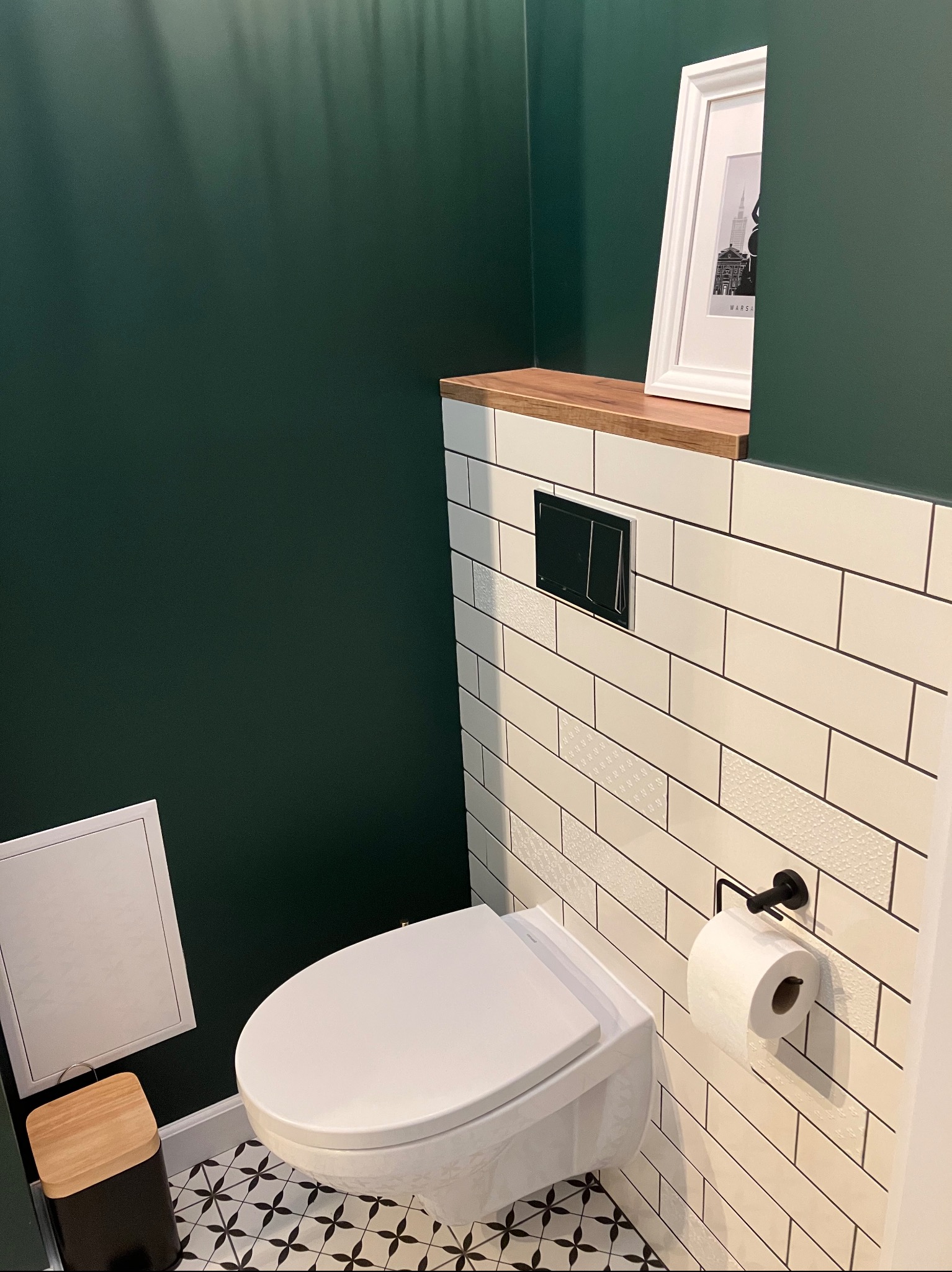 Nowoczesna łazienka z wiszącą toaletą, białą glazurą z czarną fugą i ciemnozieloną ścianą. Na półce dekoracja w ramce. Czarno-biała podłoga w geometryczny wzór.