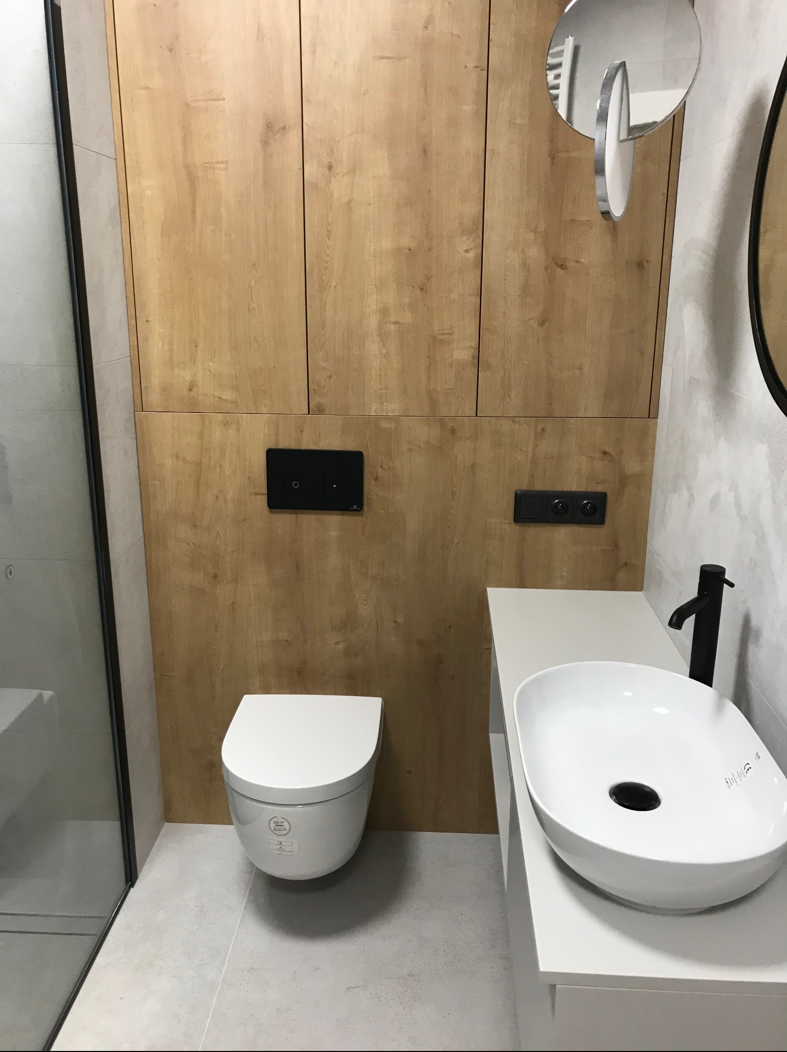 Nowoczesna łazienka po remoncie: biała umywalka nablatowa z czarną armaturą, podwieszana toaleta i drewniane szafki w zabudowie. Minimalistyczny design.