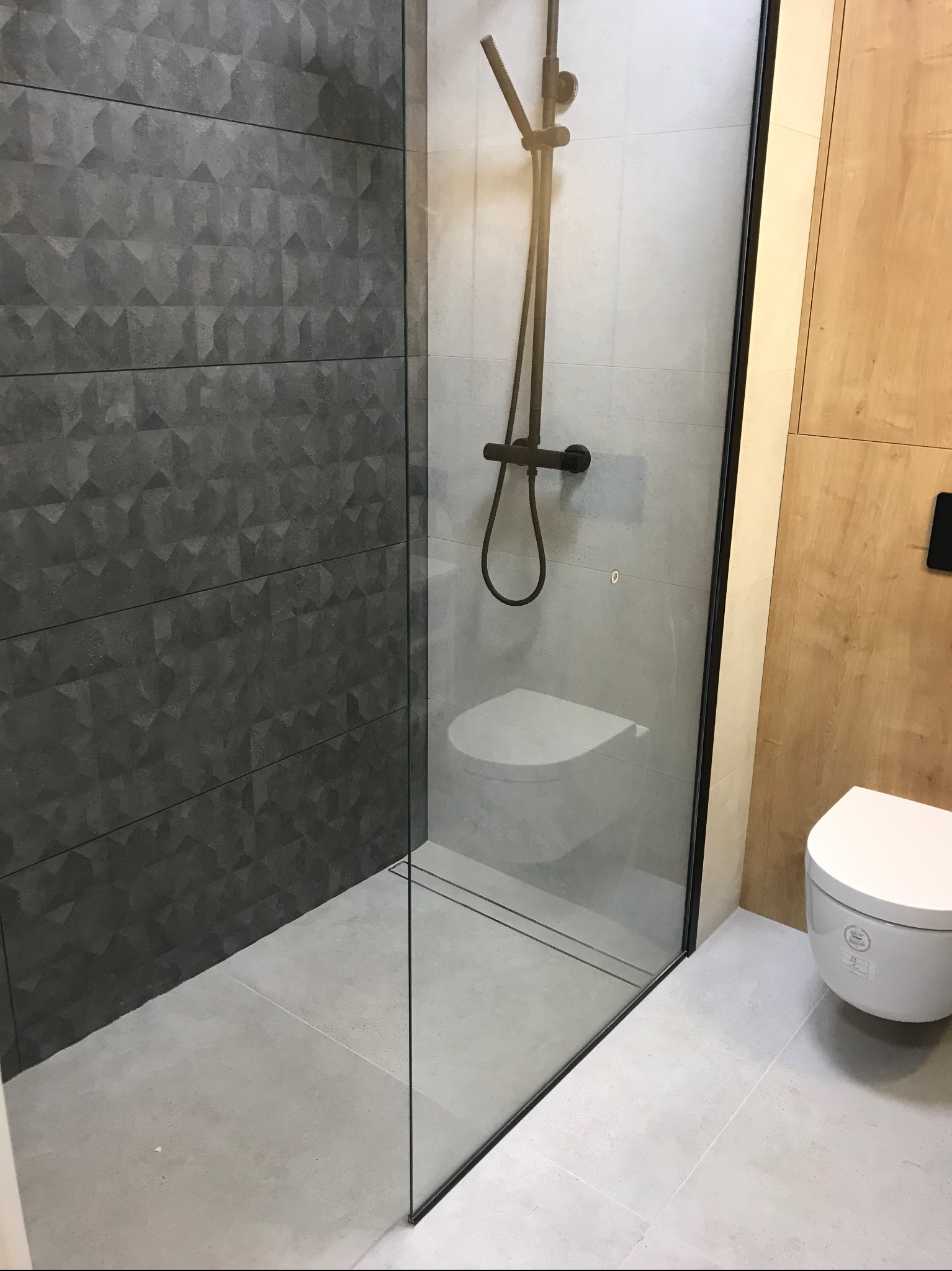 Nowoczesna kabina walk-in z czarną armaturą i geometryczną szarą okładziną ścienną, uzupełniona jasnymi płytkami podłogowymi i drewnianą zabudową.