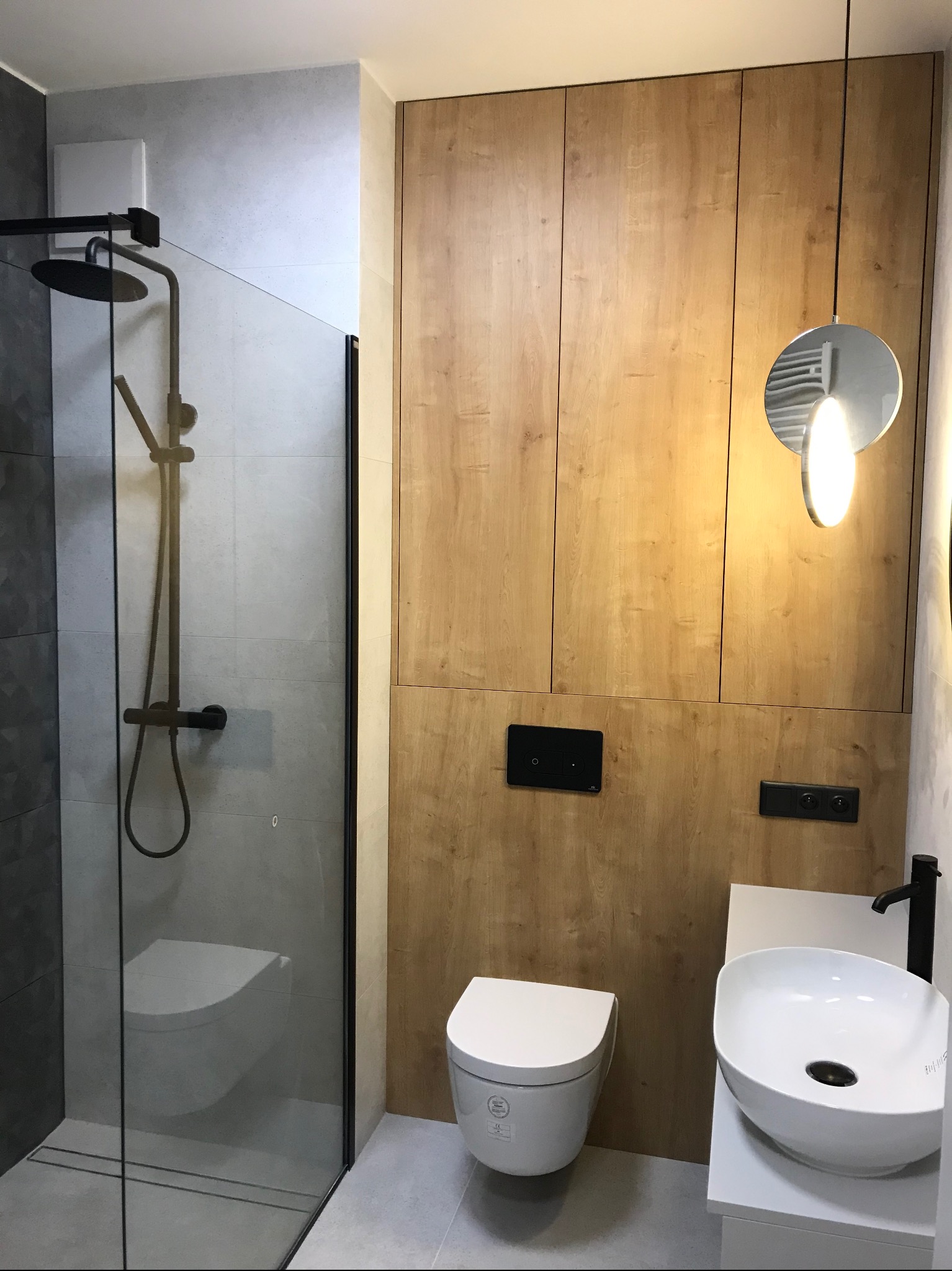 Nowoczesna łazienka z kabiną typu walk-in, drewnianą zabudową, podwieszaną toaletą i okrągłą lampą. Minimalistyczny design z czarnymi akcentami.