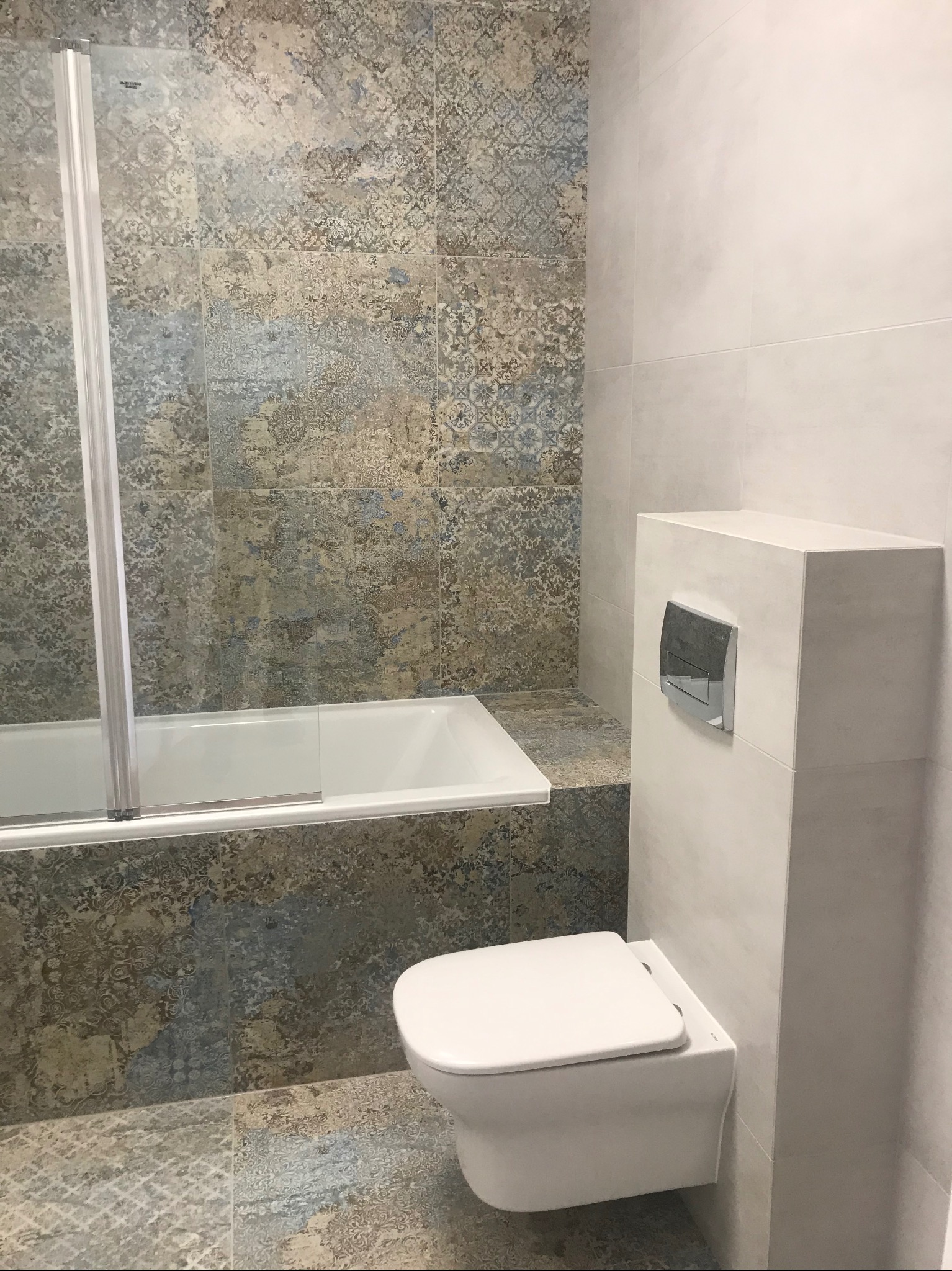 Nowoczesna łazienka z wanną za szklaną ścianką i podwieszaną toaletą. Płytki w odcieniach beżu i błękitu z delikatnym wzorem. Minimalistyczny design.