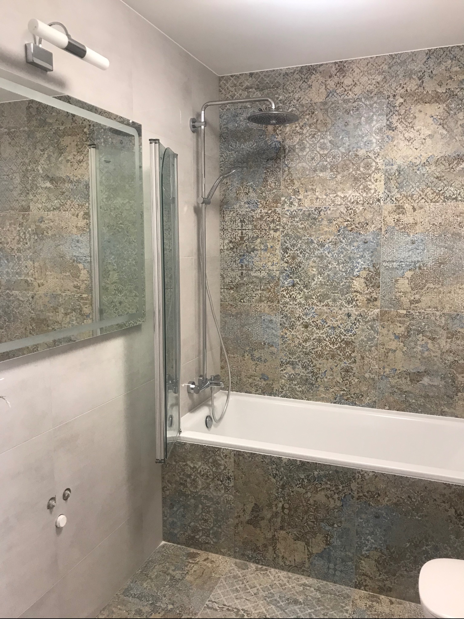 Wykończona łazienka z wanną obudowaną dekoracyjnymi płytkami, szklanym parawanem i deszczownicą. Lustro z oświetleniem nad umywalką. Płytki podłogowe w odcieniach szarości.