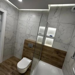 DONG BAU GROUP SPÓŁKA Z OGRANICZONĄ ODPOWIEDZIALNOŚCIĄ - Nowoczesna łazienka po remoncie: białe marmurowe płytki, drewnopodobna podłoga, podświetlane wnęki, minimalistyczna toaleta i szklany parawan nawannowy.